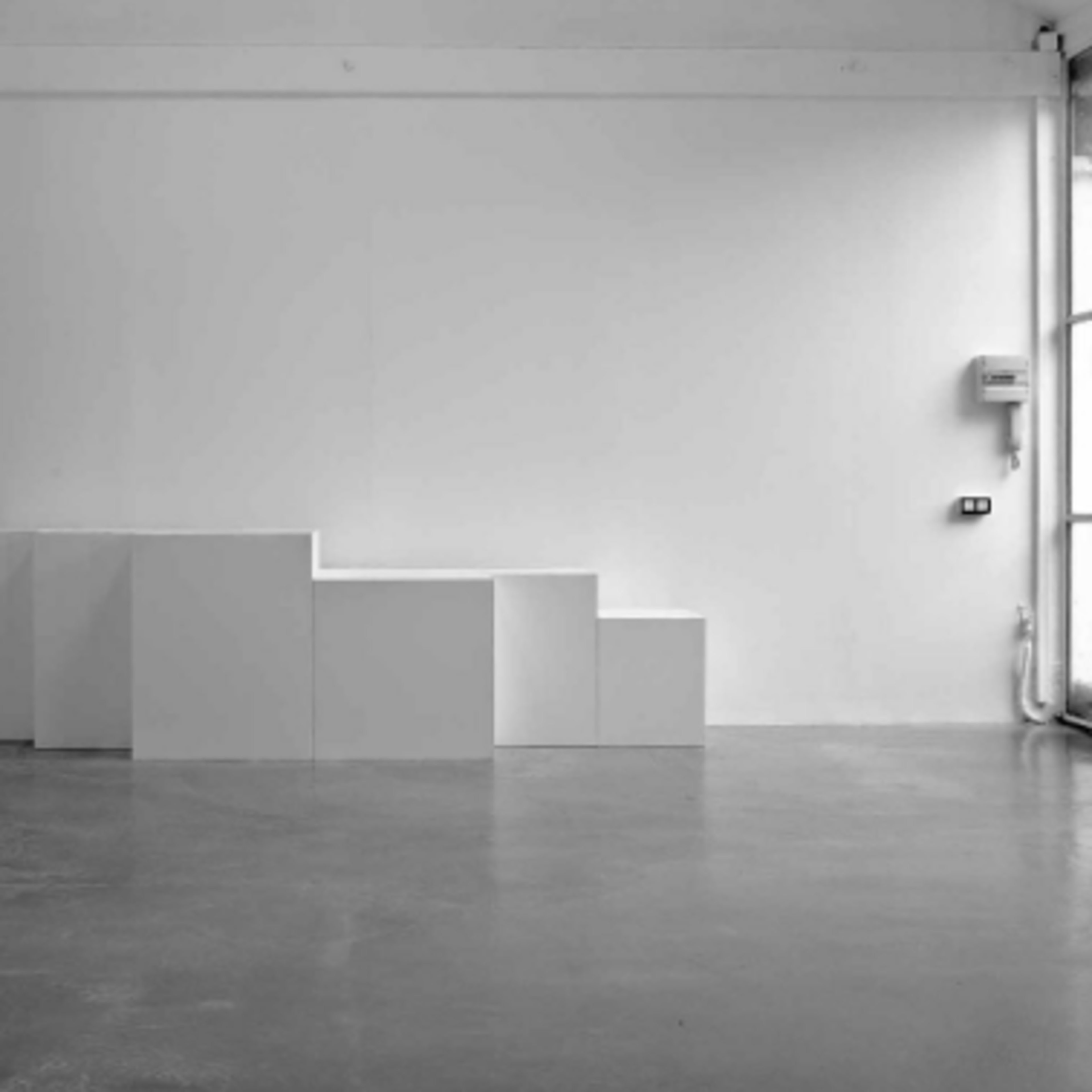 A minimalist gallery space featuring a series of white, stepped cuboid structures against a plain white wall.