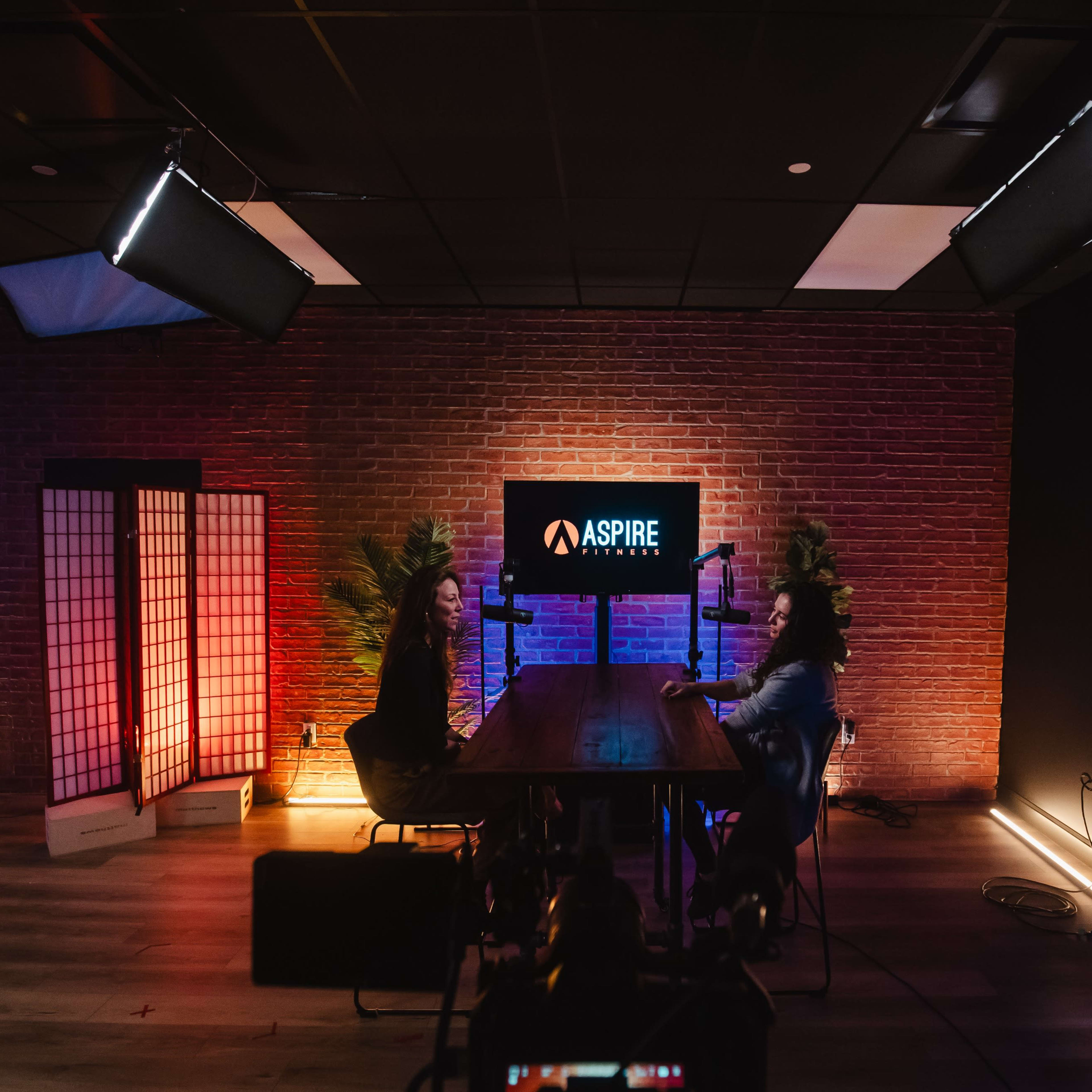 A well-lit studio setting features two individuals seated at a wooden table with microphones, against a backdrop of brick walls and an "ASPIRE" sign.