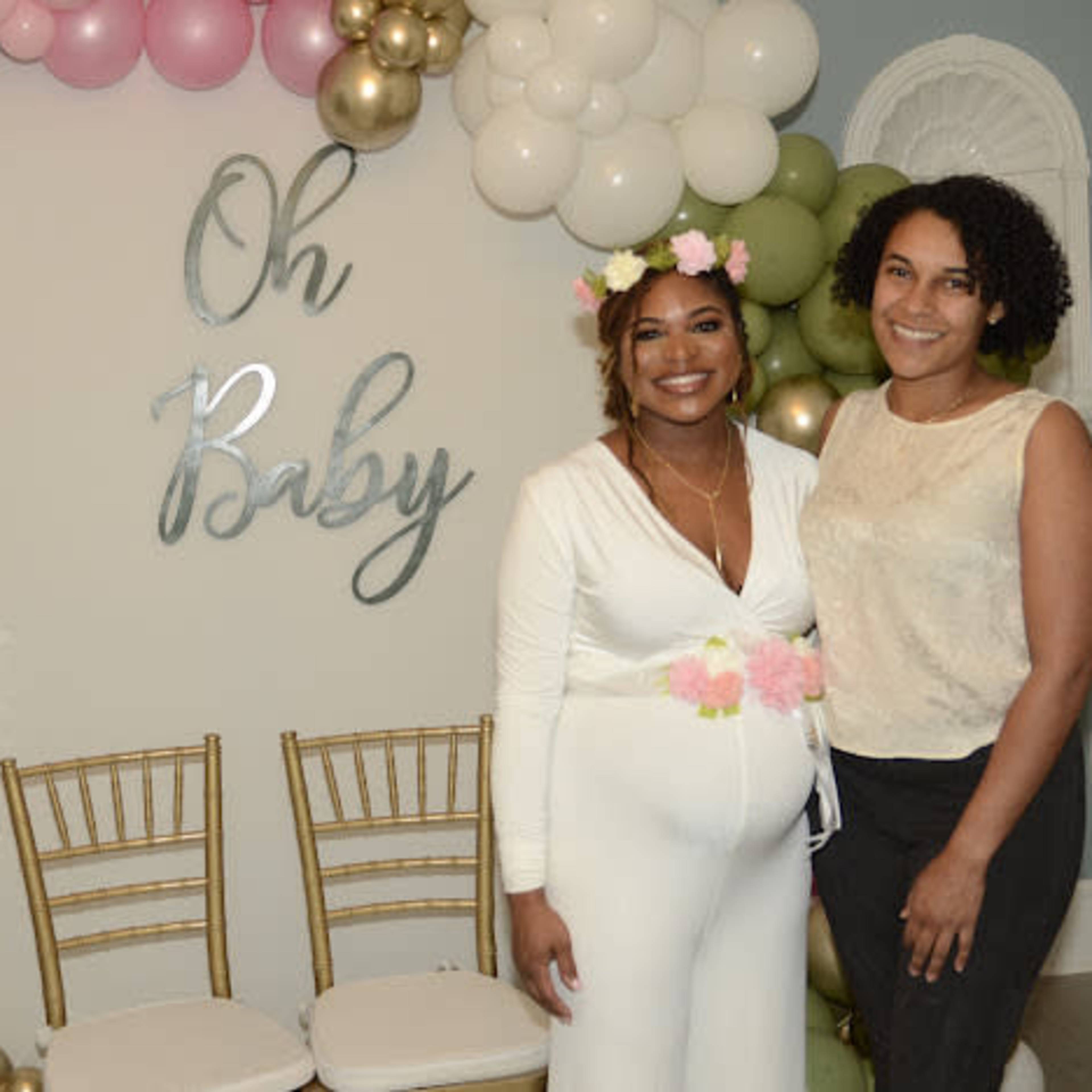 Two women stand in front of a decorated wall with the words "Oh Baby," surrounded by balloons and a display of blocks.