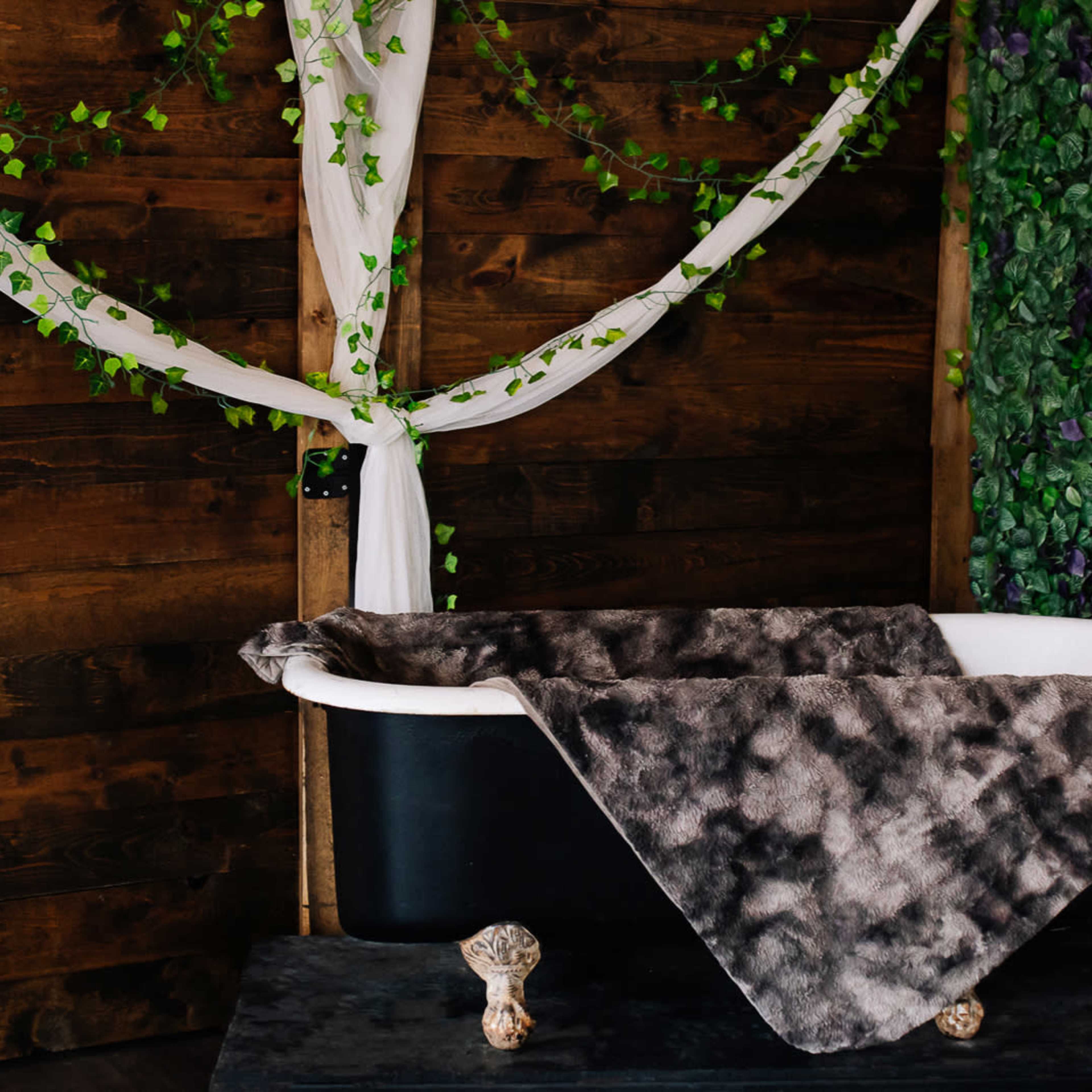 A vintage bathtub with a dark, textured blanket draped over it is set against a backdrop of wooden walls and greenery.