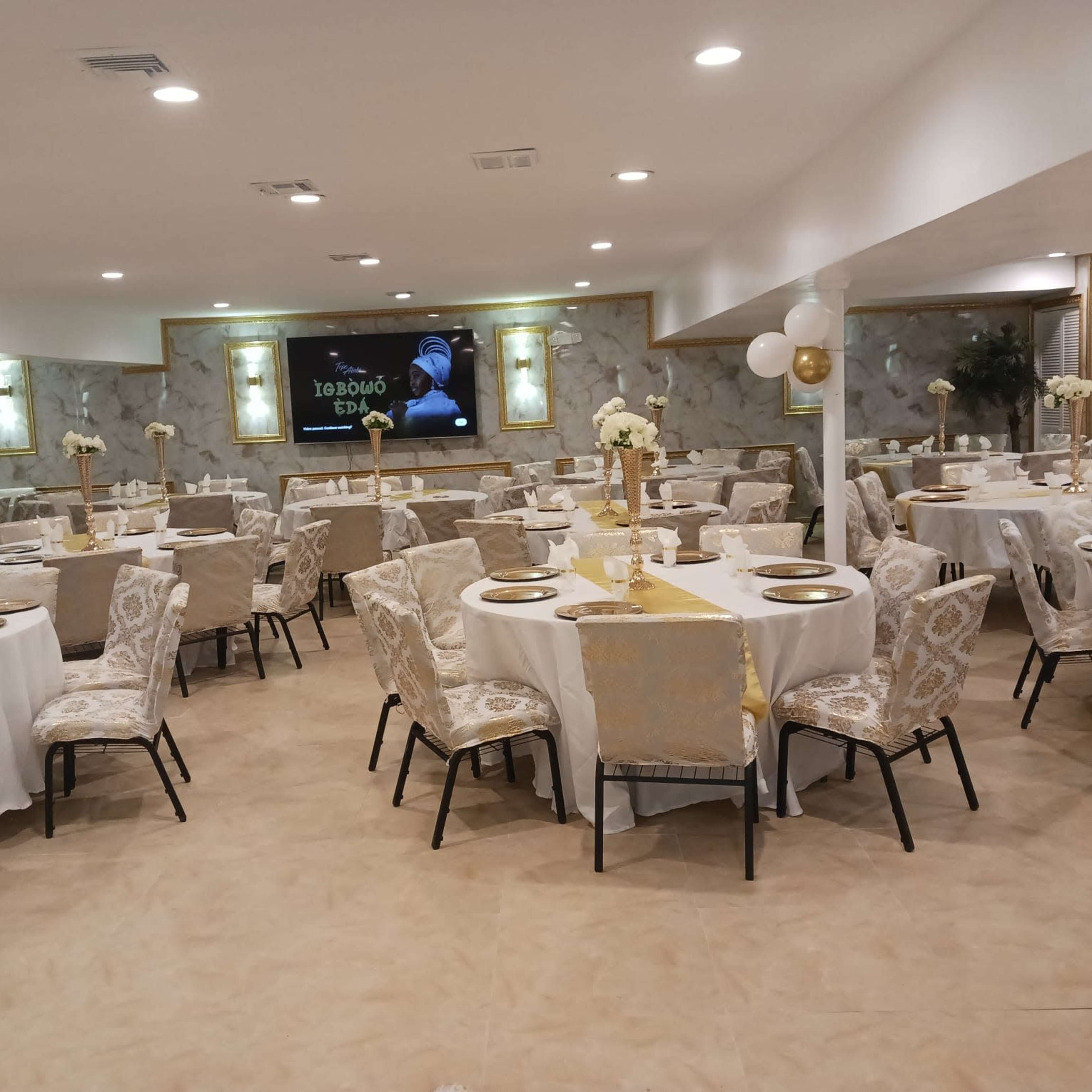 A banquet hall with round tables dressed in white tablecloths and gold accents, surrounded by upholstered chairs, and featuring wall decorations and a large screen at the front.