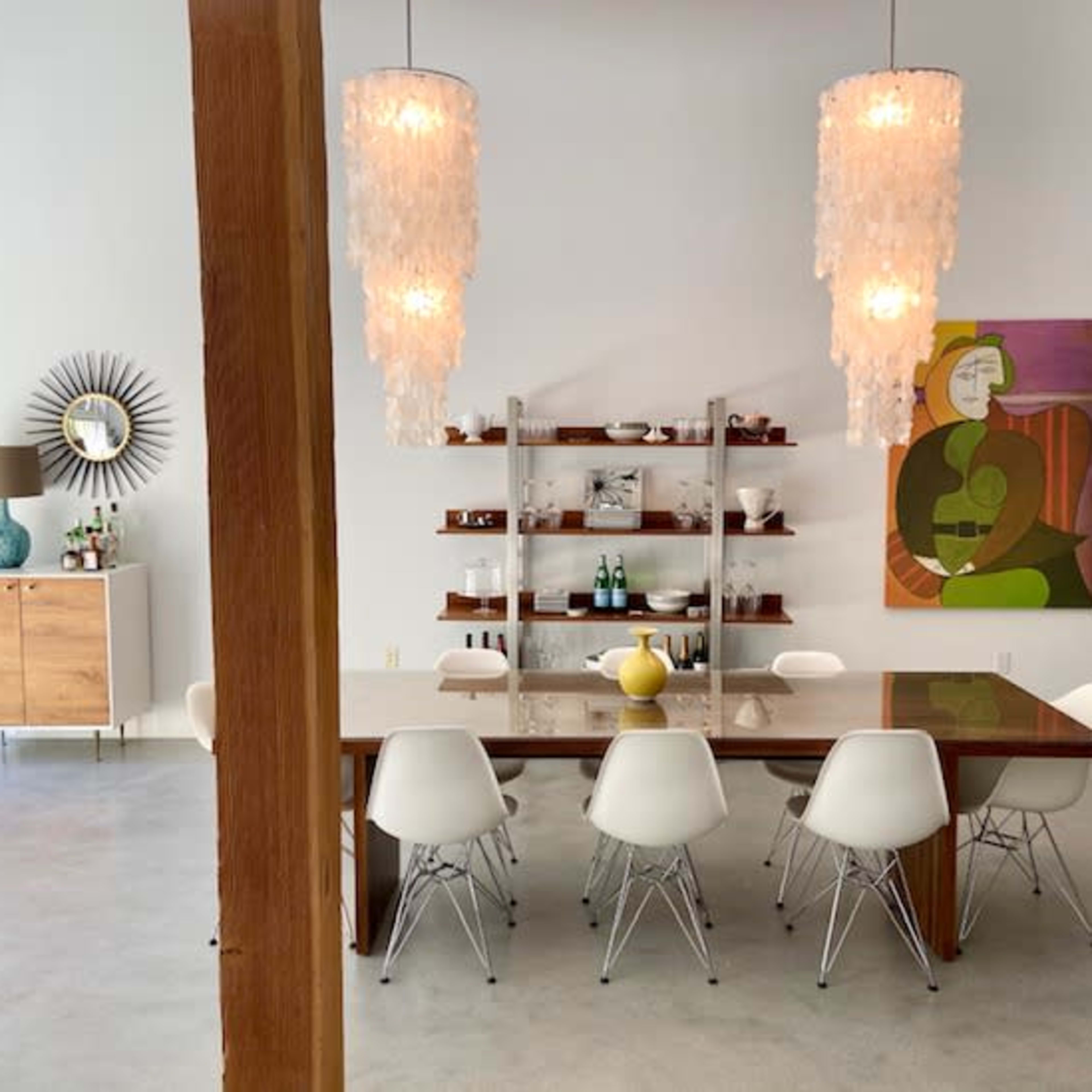 A modern dining area features a wooden table surrounded by white chairs, with two large pendant lights and a colorful wall painting in the background.