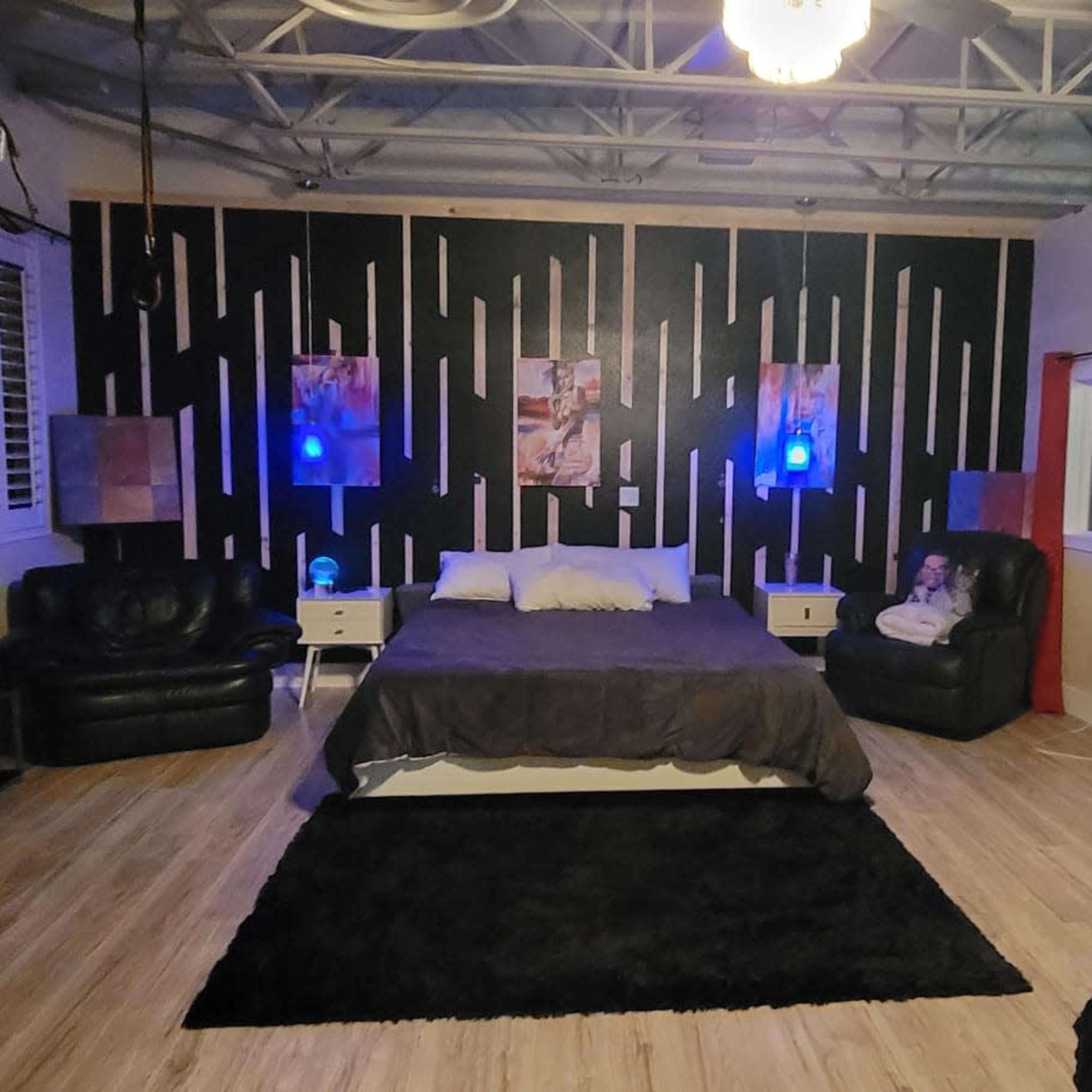A spacious room featuring a large bed in the center, dark furniture, and blue ambient lighting, with a workspace visible in the background.