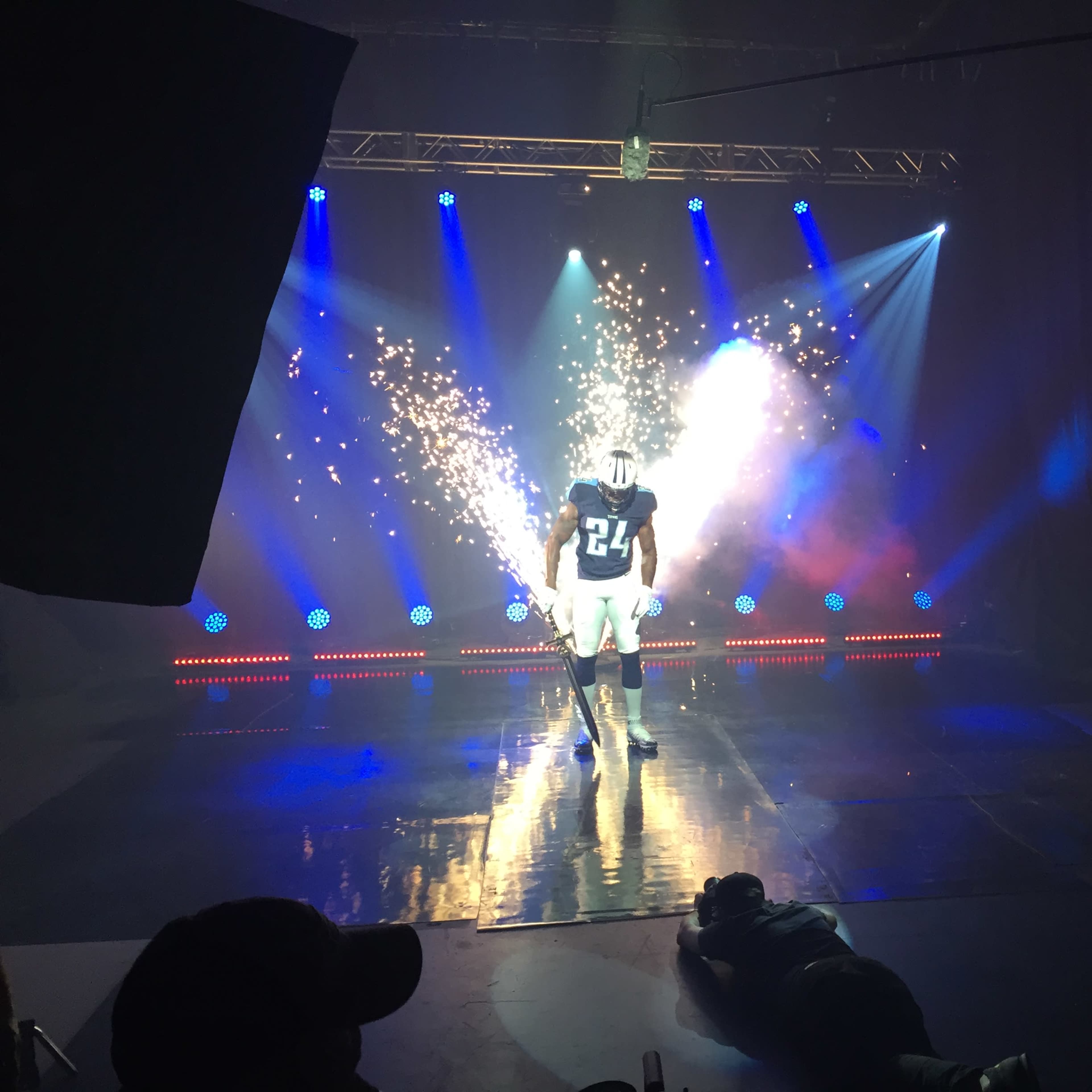A football player in a blue jersey with the number 24 stands on a reflective stage illuminated by bright lights and sparks.
