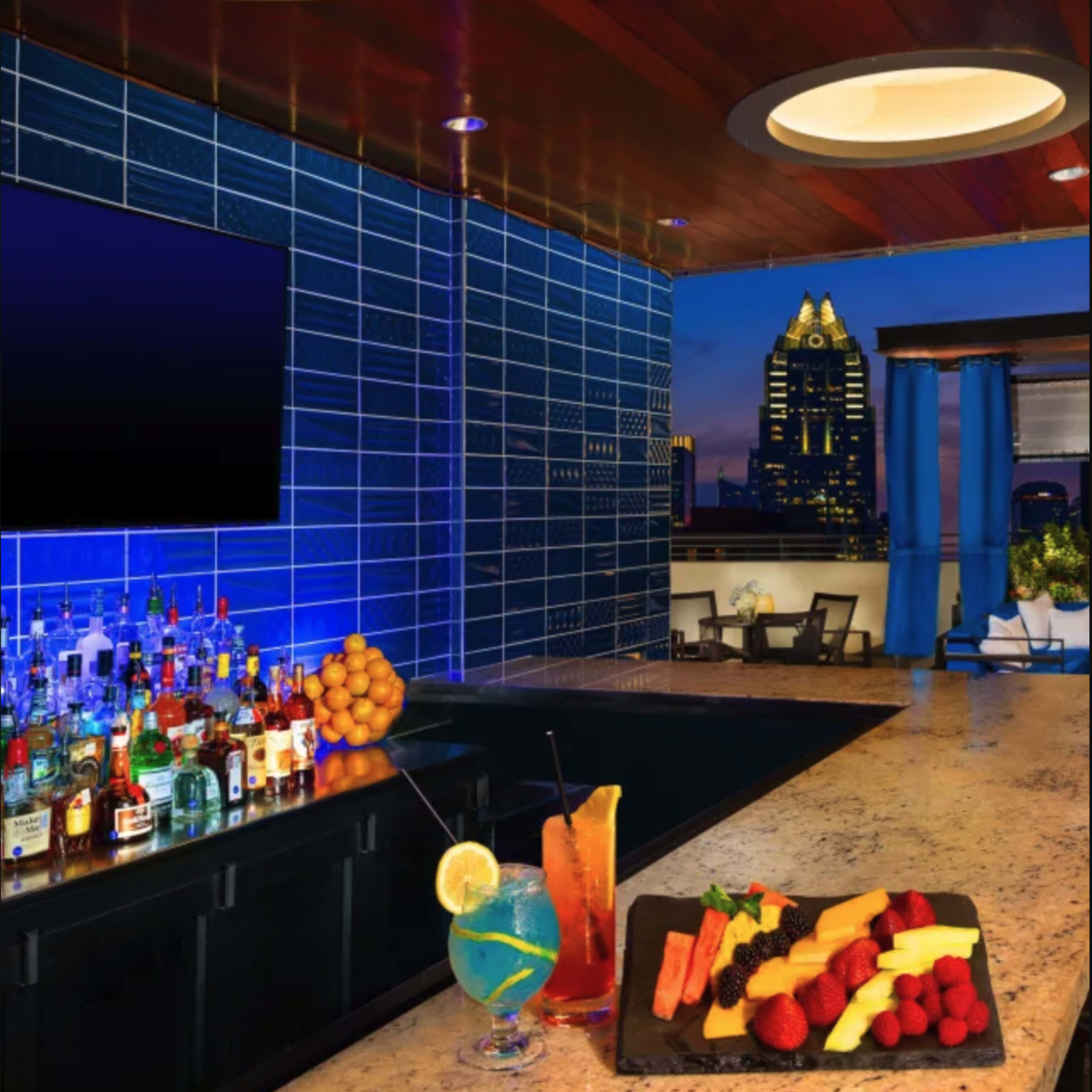 A modern bar with a blue tiled wall and a view of a city skyline features a selection of drinks and a platter of fresh fruits.