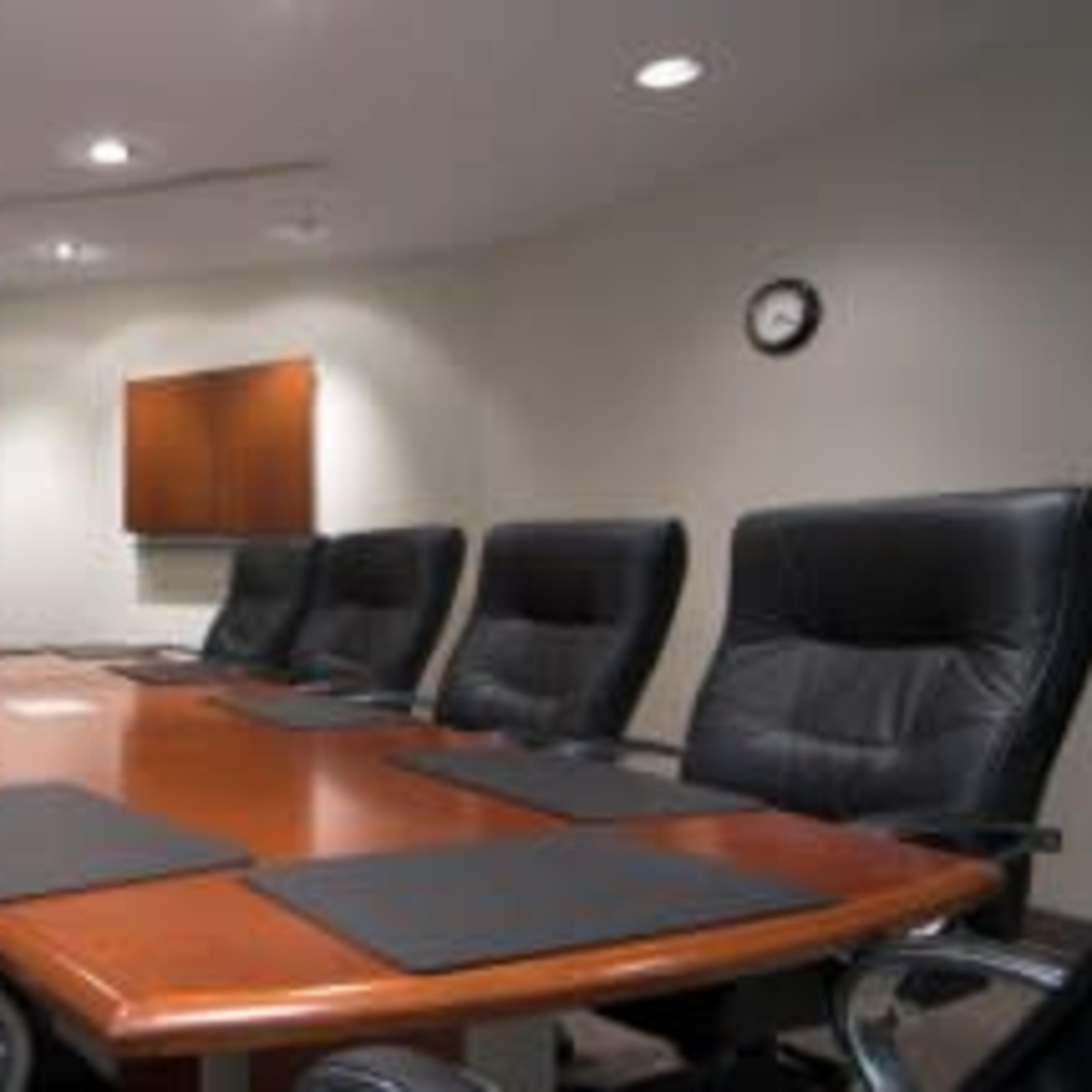 A large conference room features a wooden table surrounded by eight black leather chairs, with a clock and a television mounted on the wall.