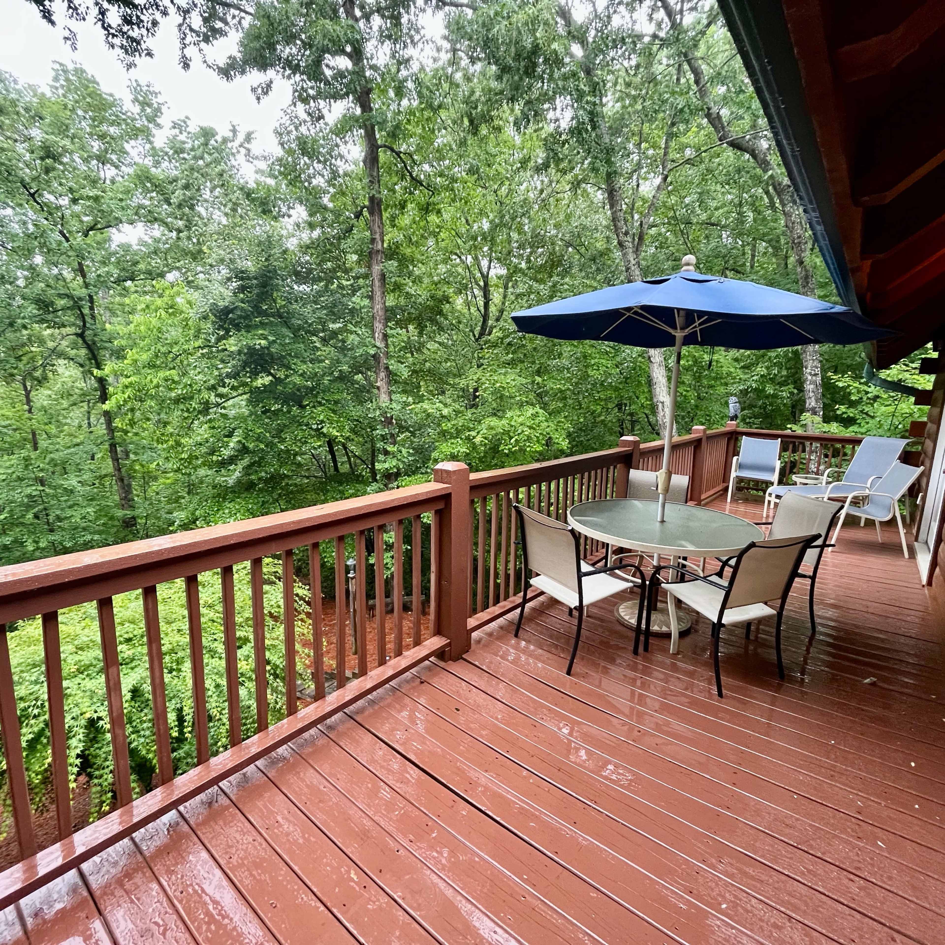 A wooden deck with a round table and chairs, accompanied by a blue umbrella, overlooks a forested area.