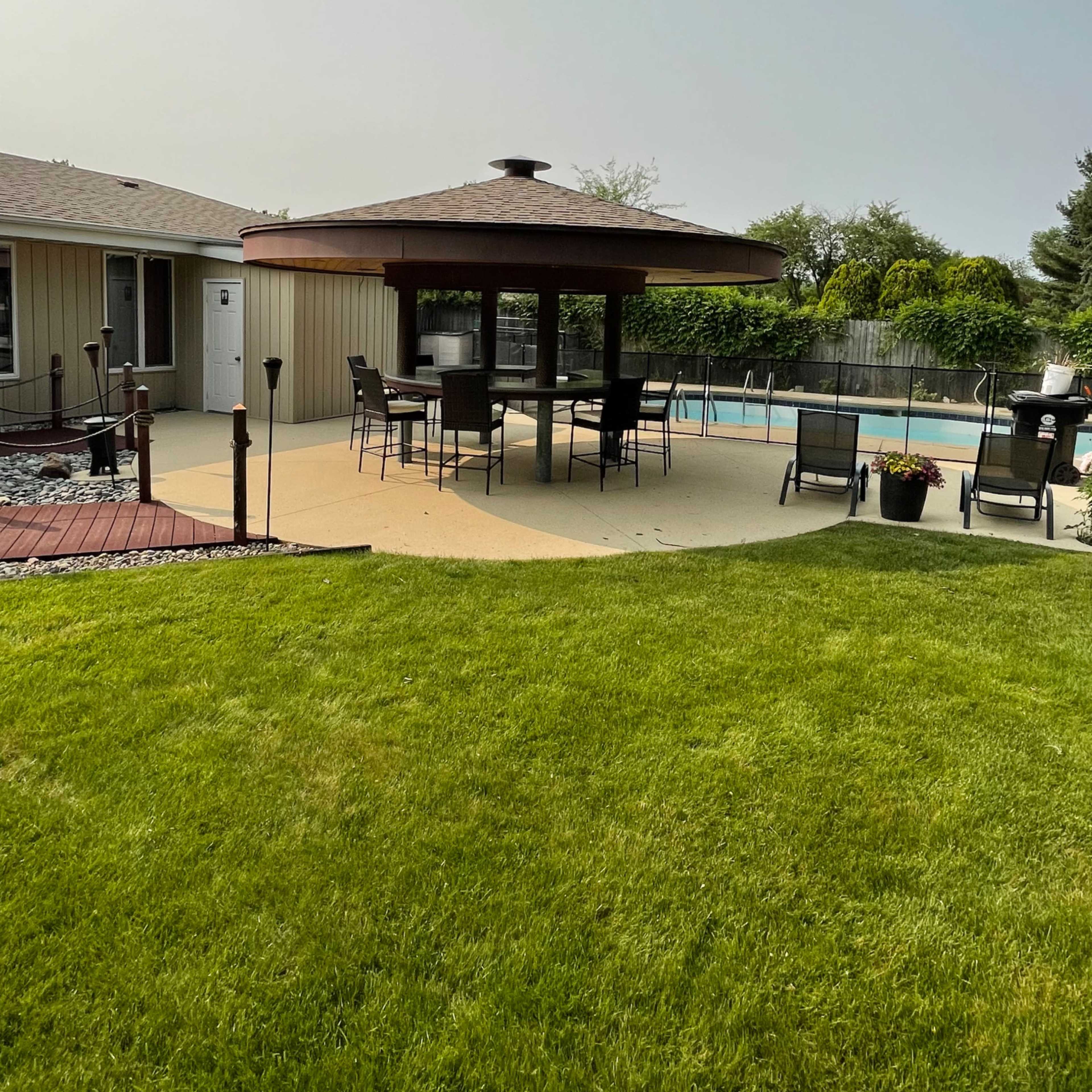 A backyard with a gazebo containing a dining set, surrounding a swimming pool and grassy area.