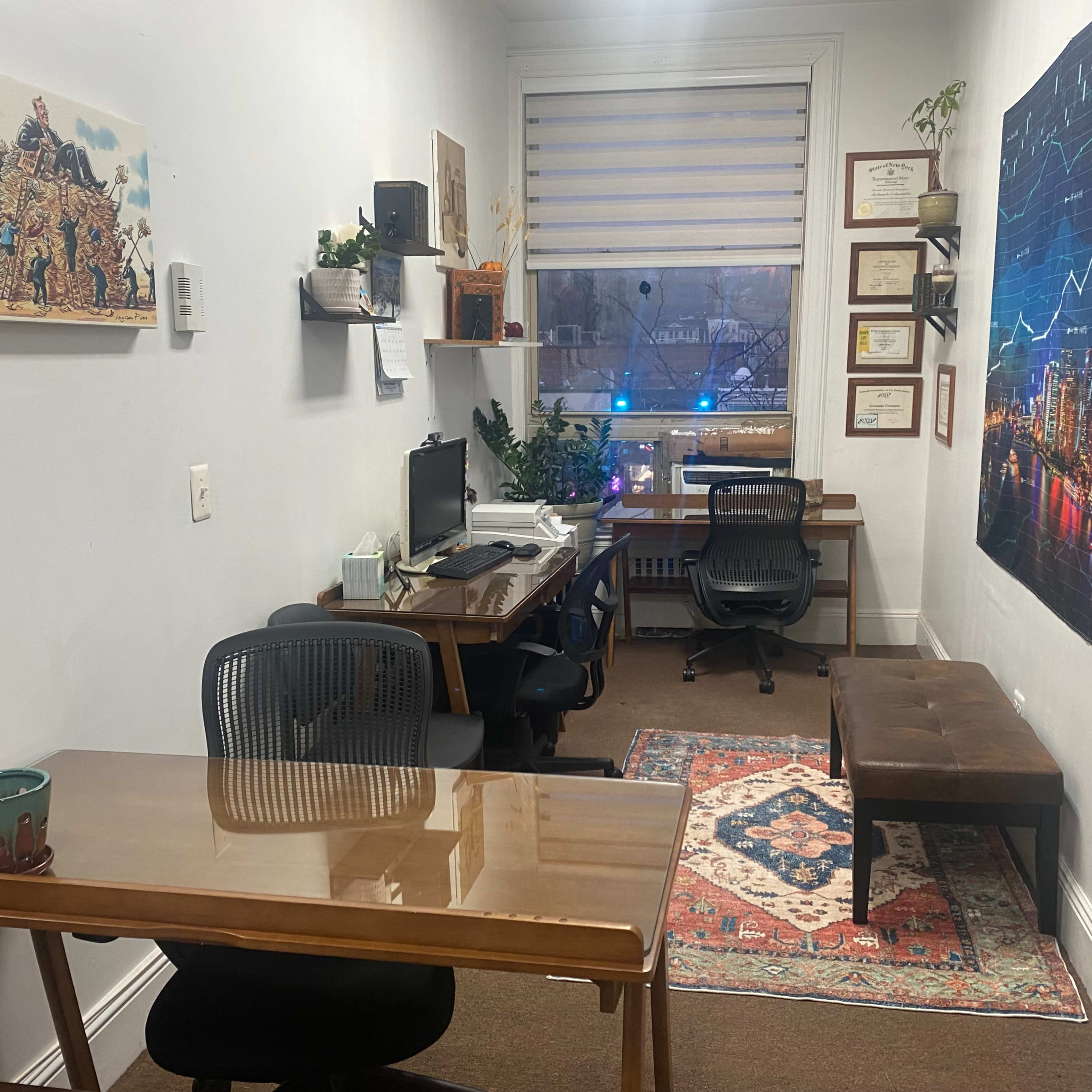 A small office space featuring a desk with a computer, a bench, a wall adorned with framed certificates, and a large graphic of a city skyline with financial charts.