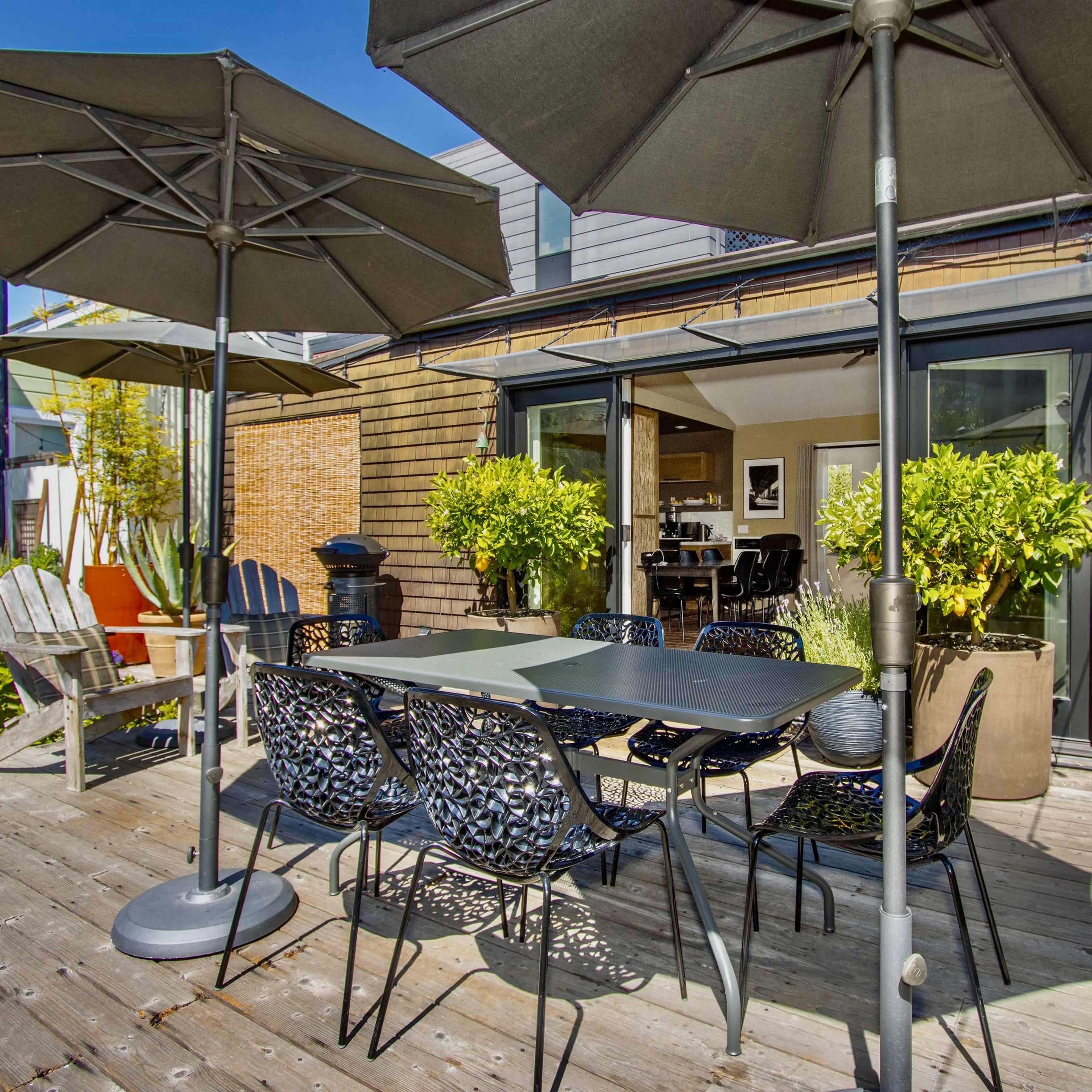 A patio area features a table with chairs and two large umbrellas, surrounded by potted plants and wooden decking.