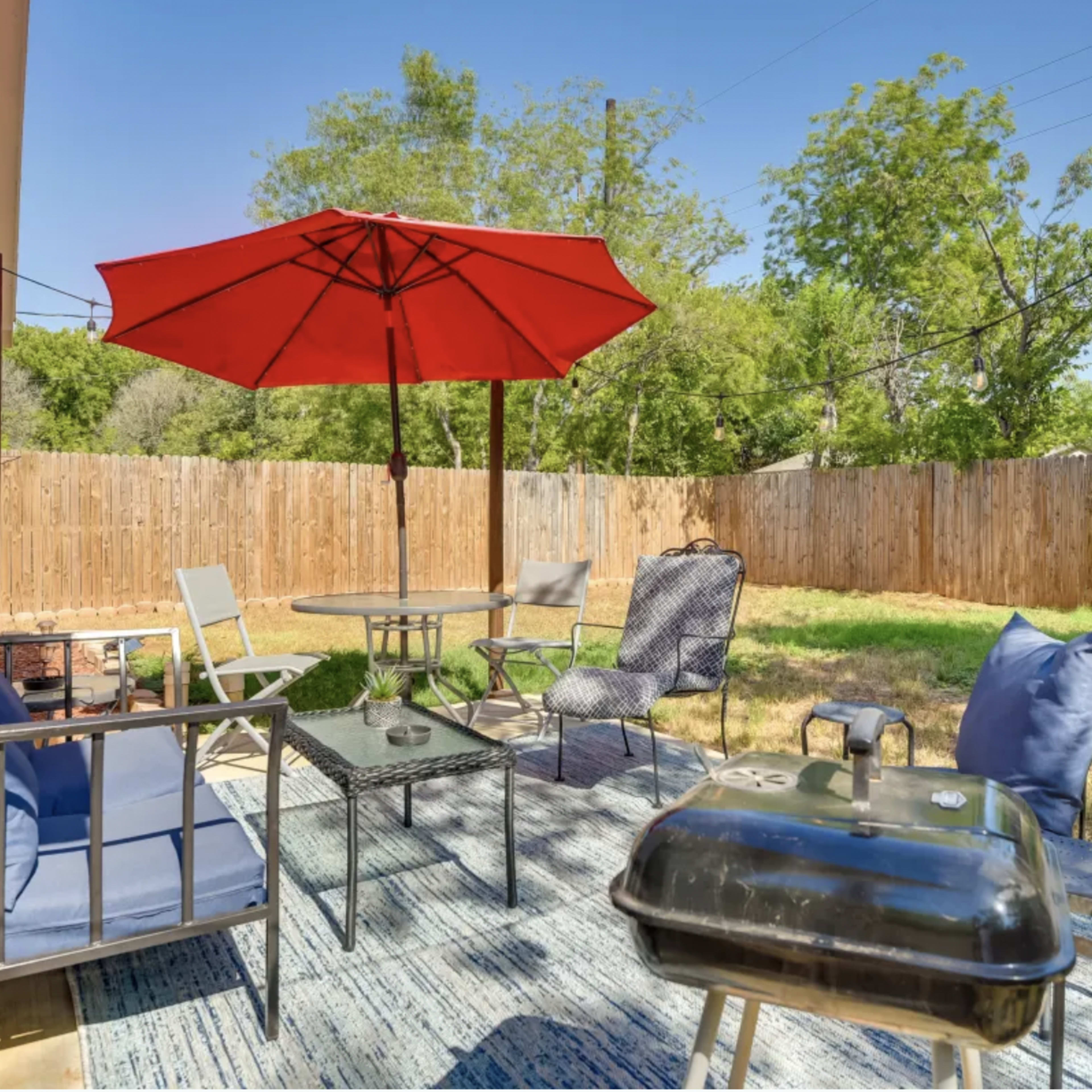 A patio area features a red umbrella over a circular table surrounded by chairs, with a charcoal grill and a grassy yard enclosed by a wooden fence.