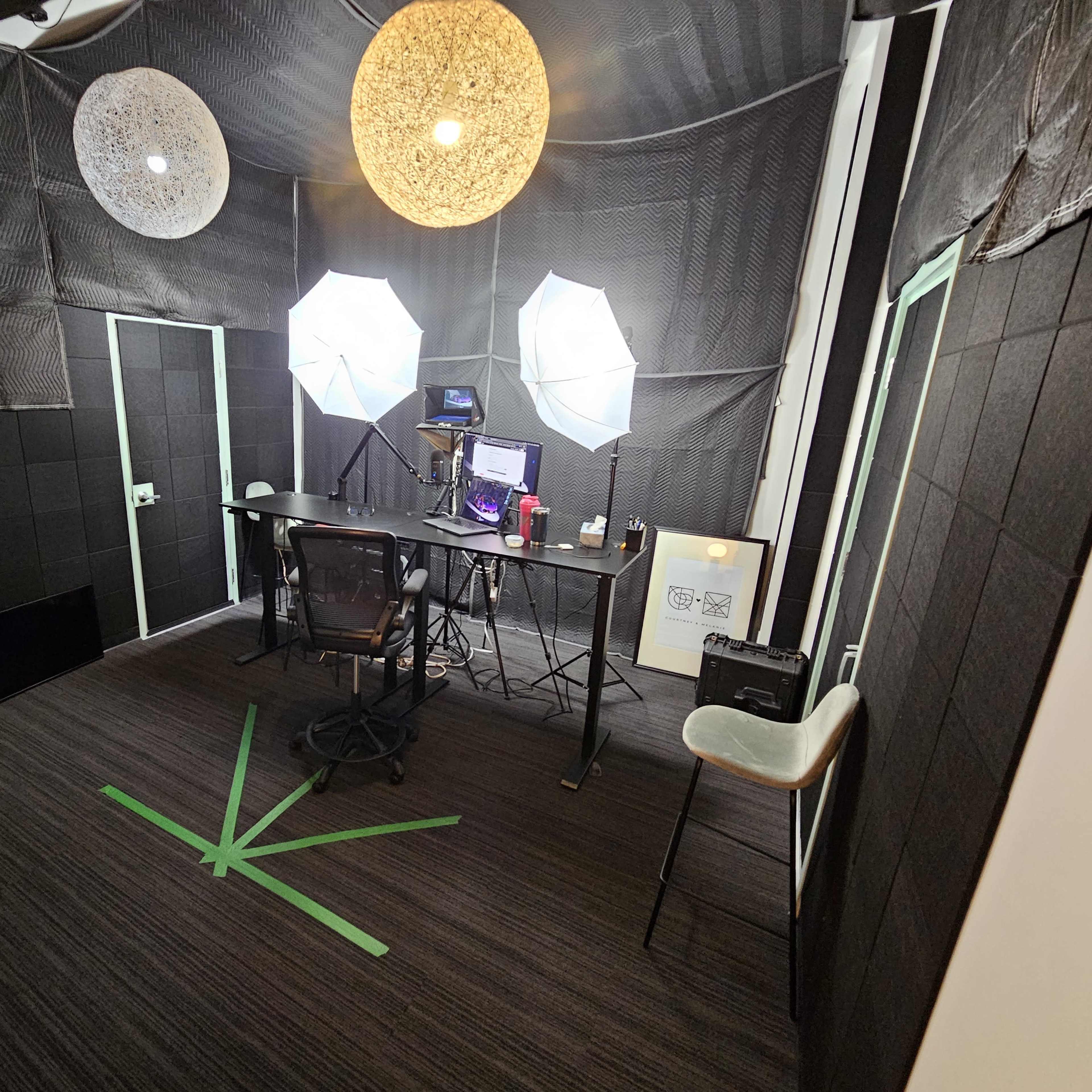 A small studio space features a desk with a computer and equipment, flanked by large softbox lights and decorated with hanging circular lamps.