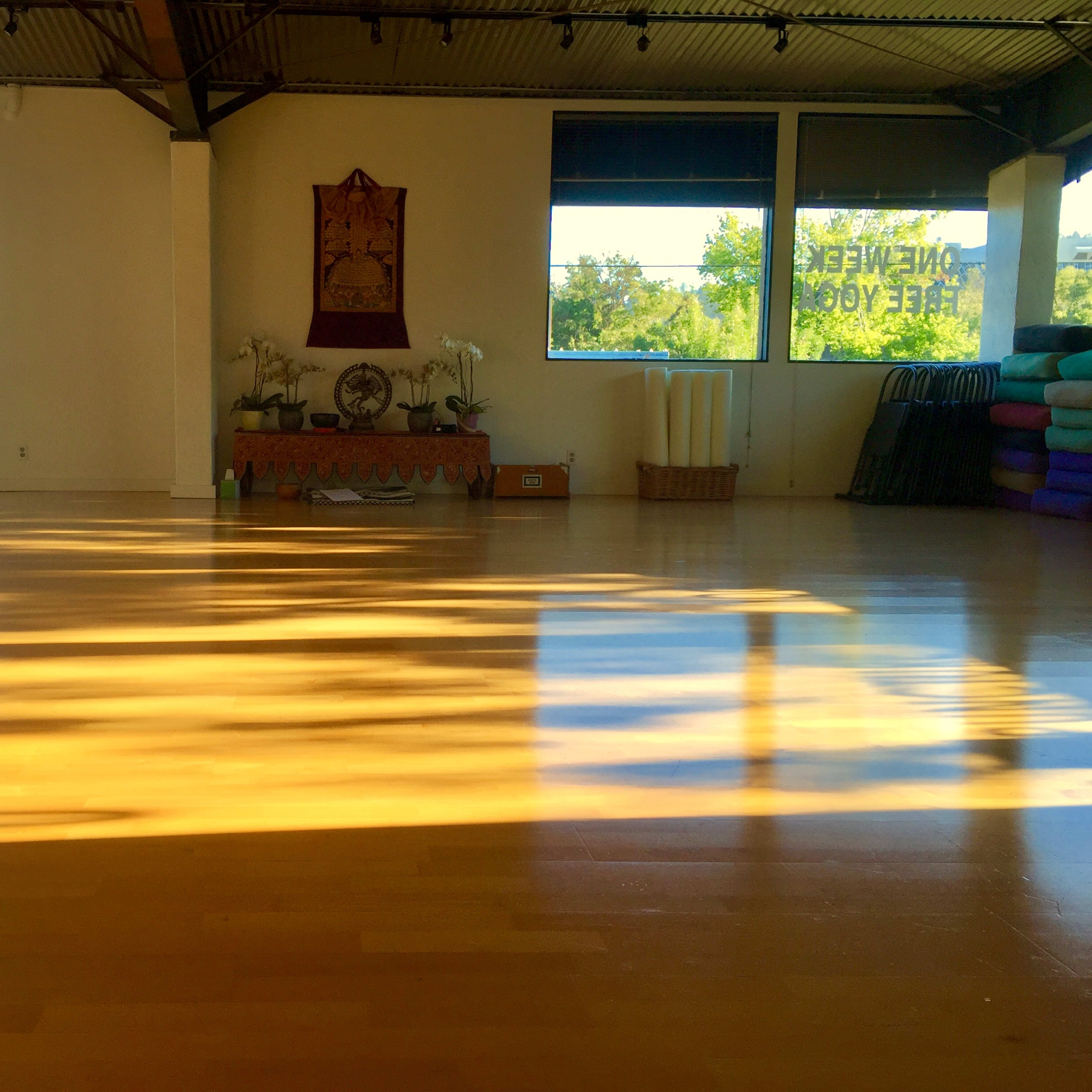 A spacious yoga studio with wooden floors, large windows, and yoga props stacked against the wall.
