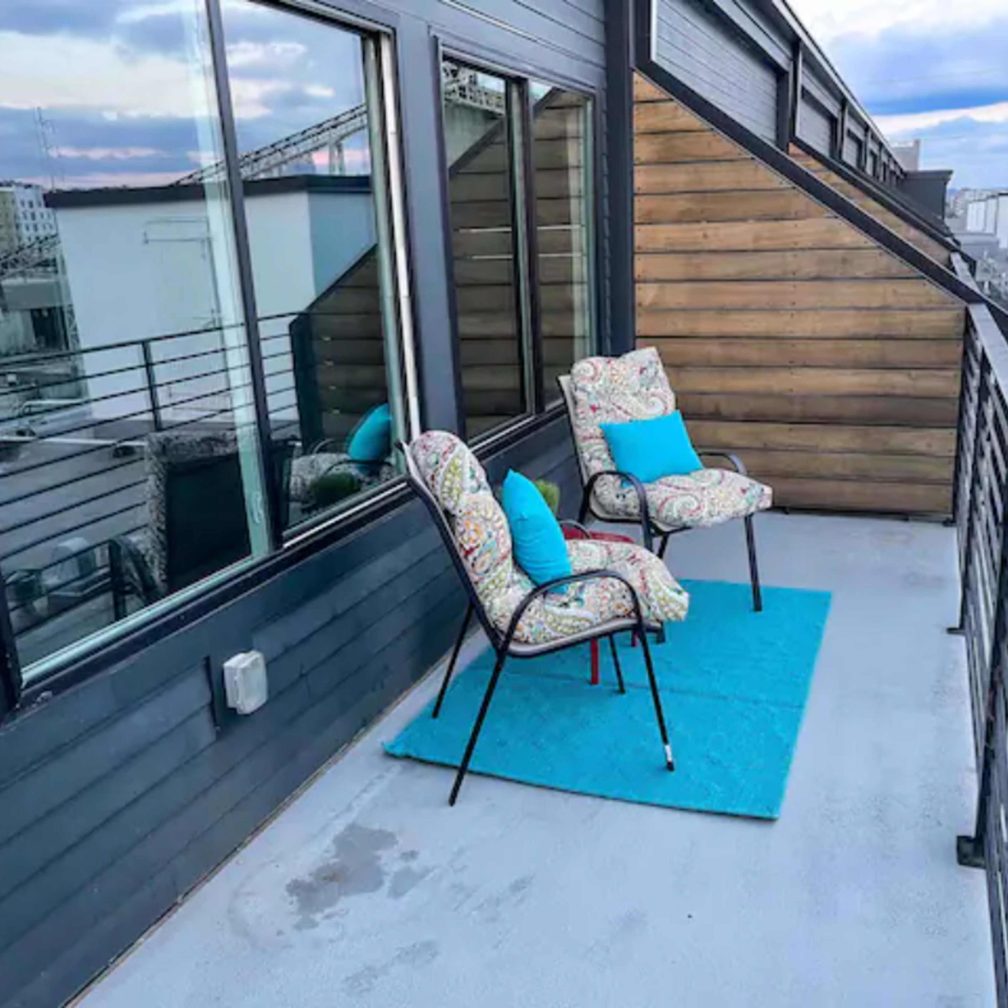 A balcony with two patterned chairs and a blue cushion on a rug, set against a backdrop of modern buildings.