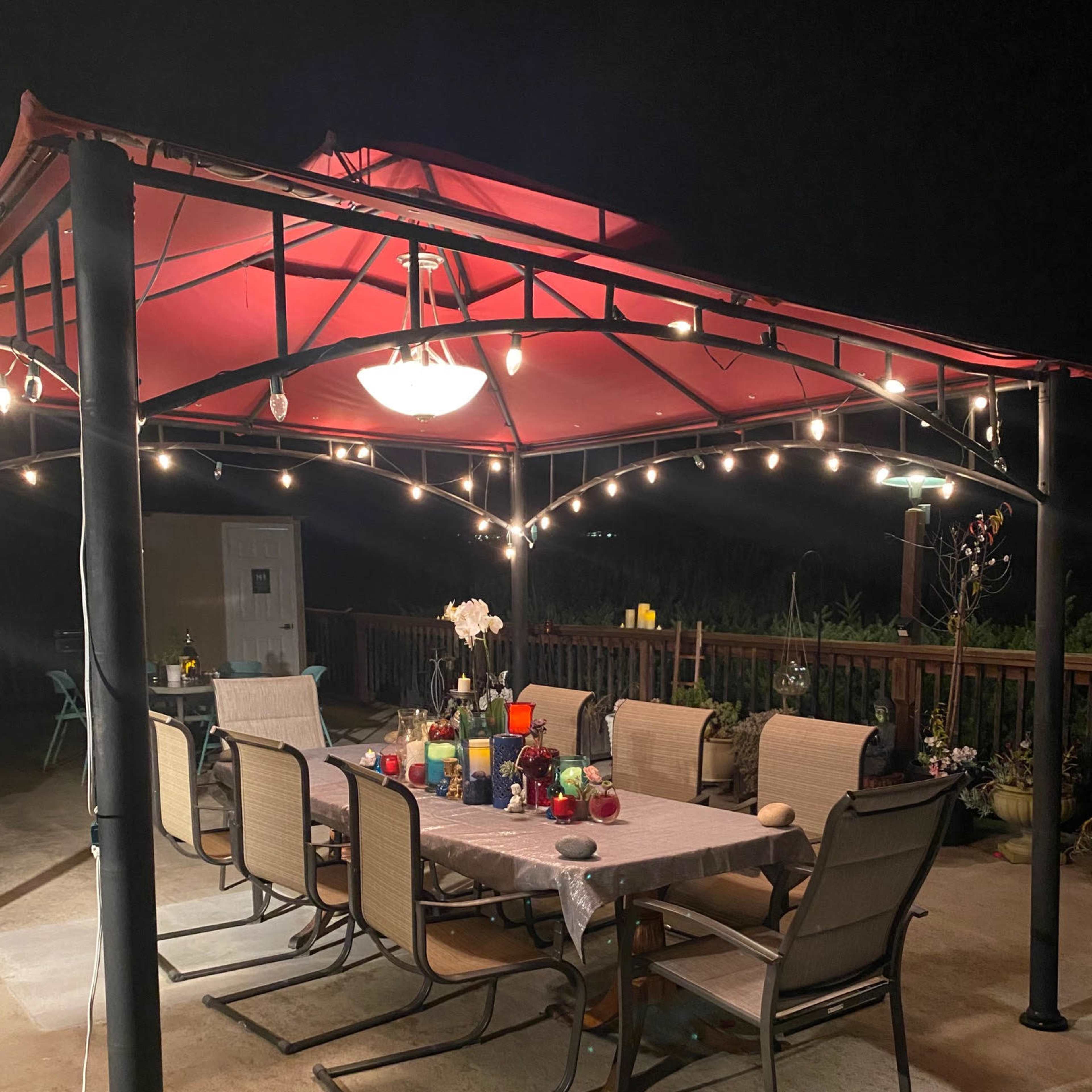 A patio with a red canopy features a table set with various drinks and surrounded by chairs, illuminated by string lights.