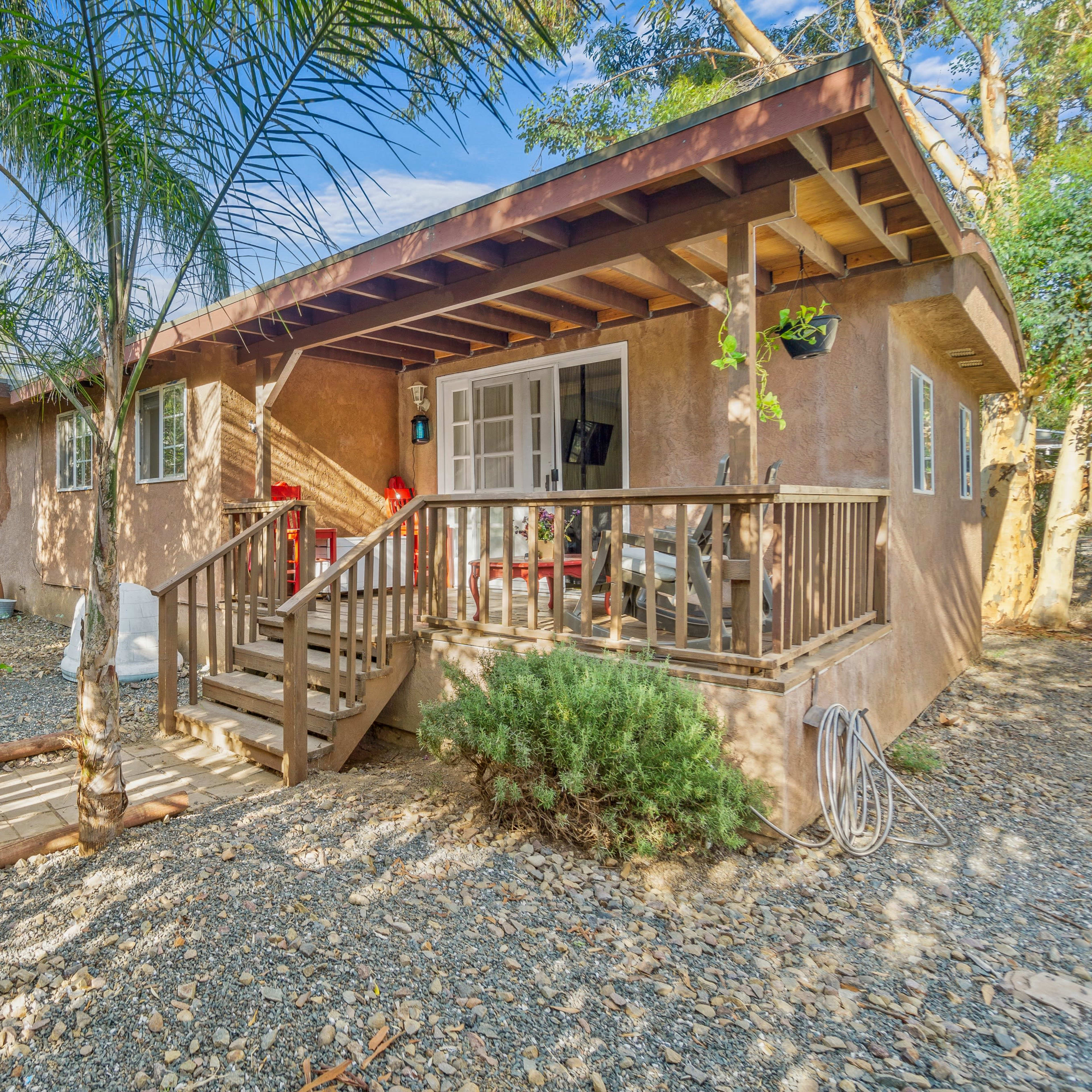A small, single-story house with a wooden porch is surrounded by palm trees and gravel landscaping.