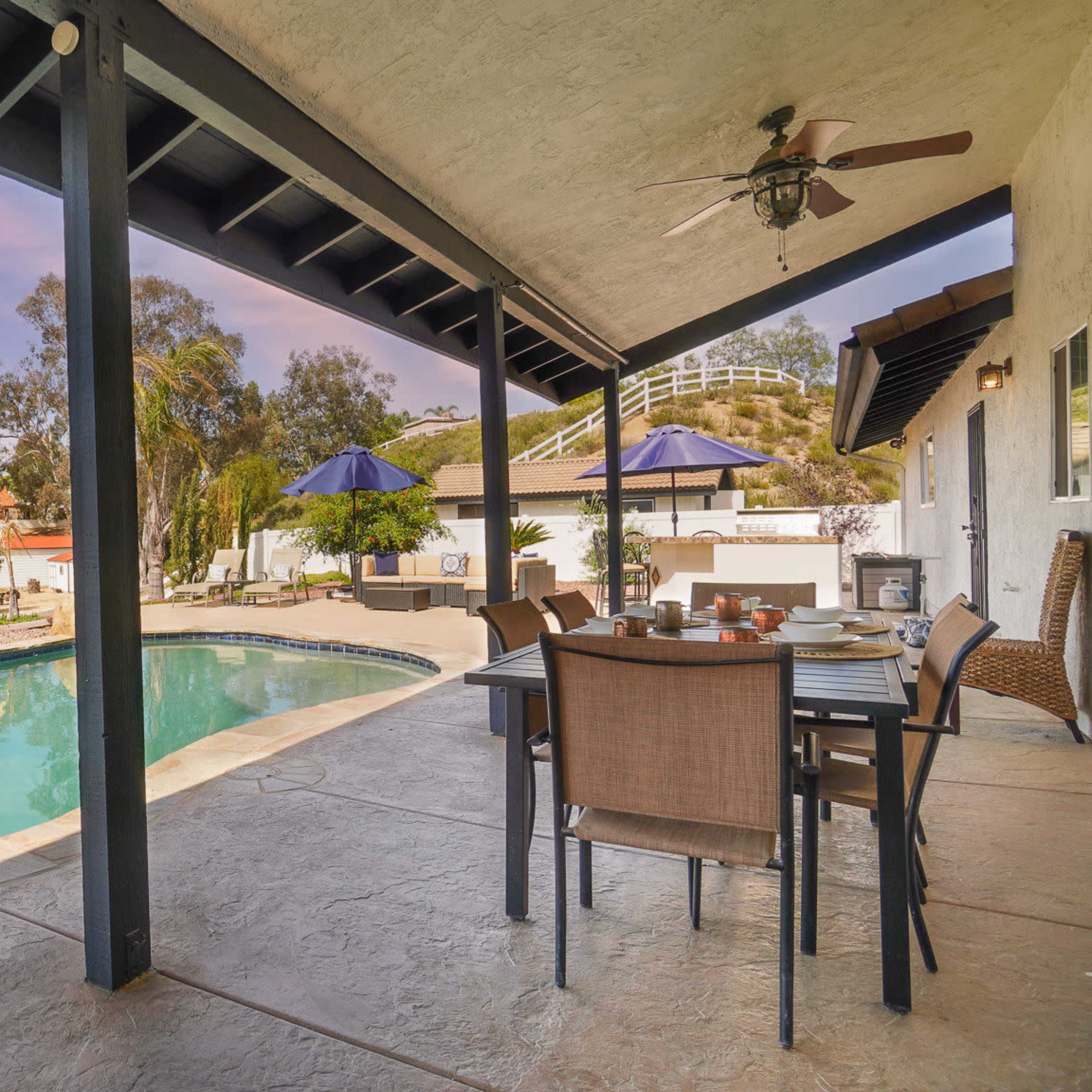 A patio with a dining table overlooks a swimming pool and landscaped yard under a colorful sky.