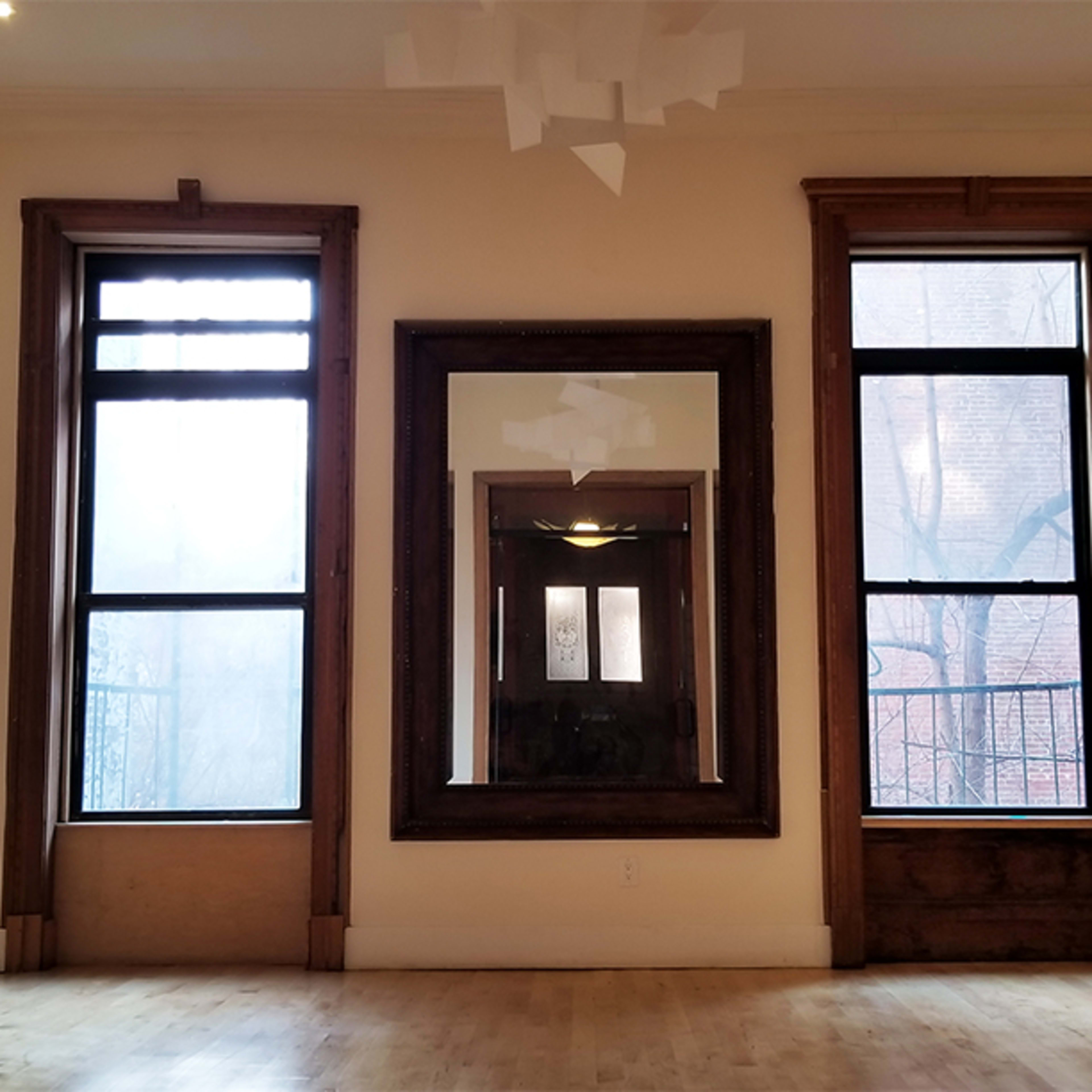A spacious, empty room with three windows and a large mirror framed in wood, highlighting the wooden trim and light-colored walls.