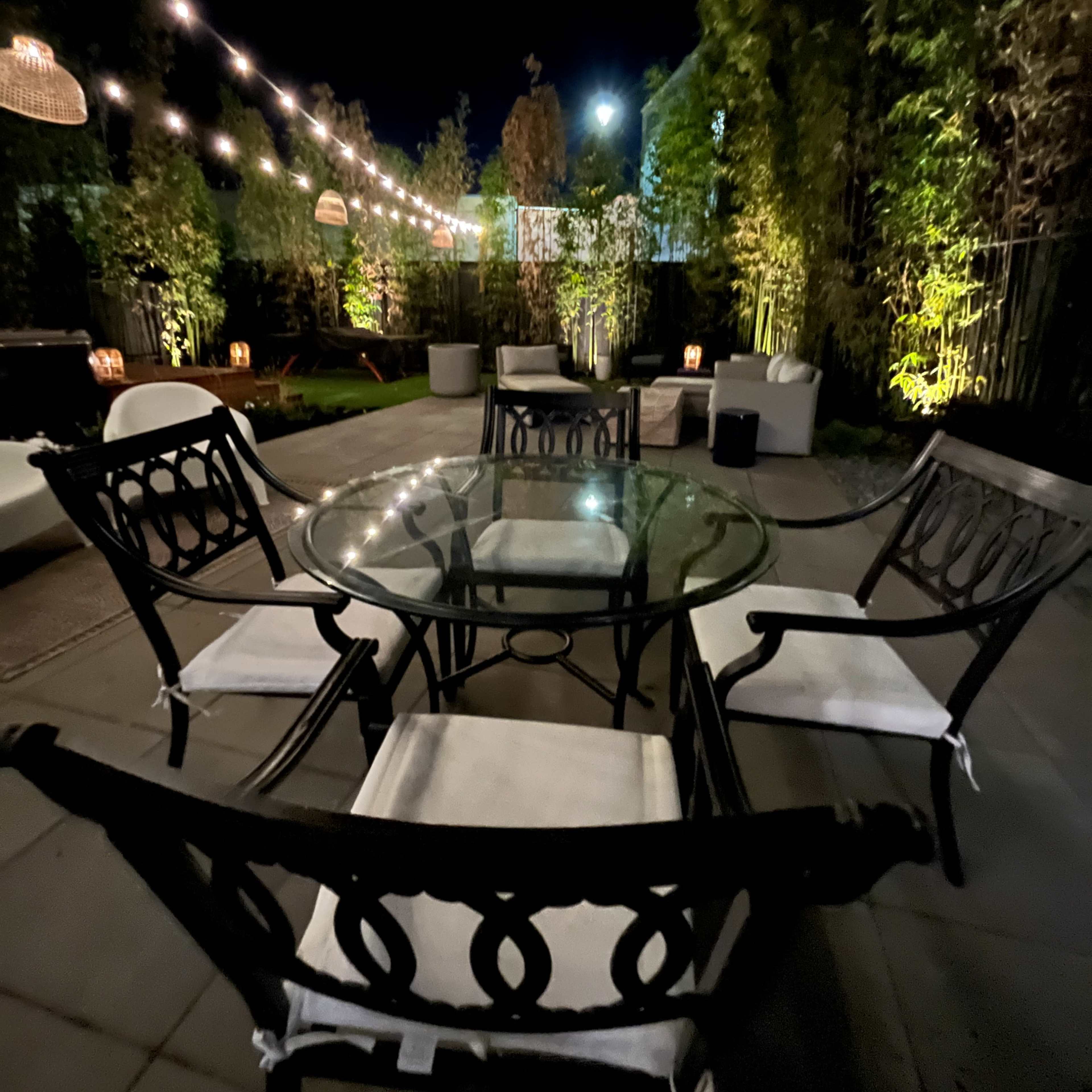 A glass-top table surrounded by black metal chairs is illuminated by string lights in a landscaped outdoor patio at night.