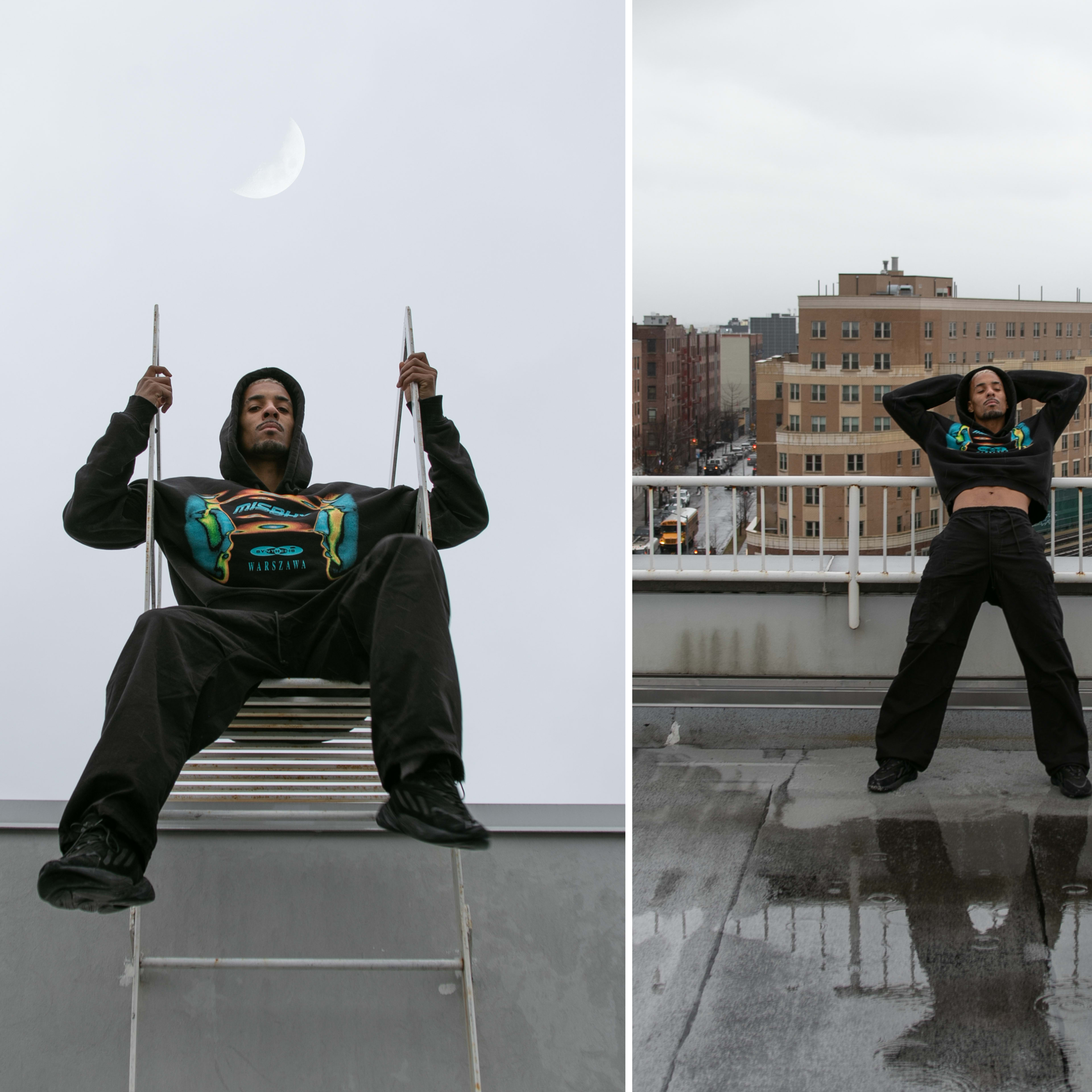A person sits on a metal ladder against a gray sky, while another person poses with arms raised on a rooftop with buildings in the background.