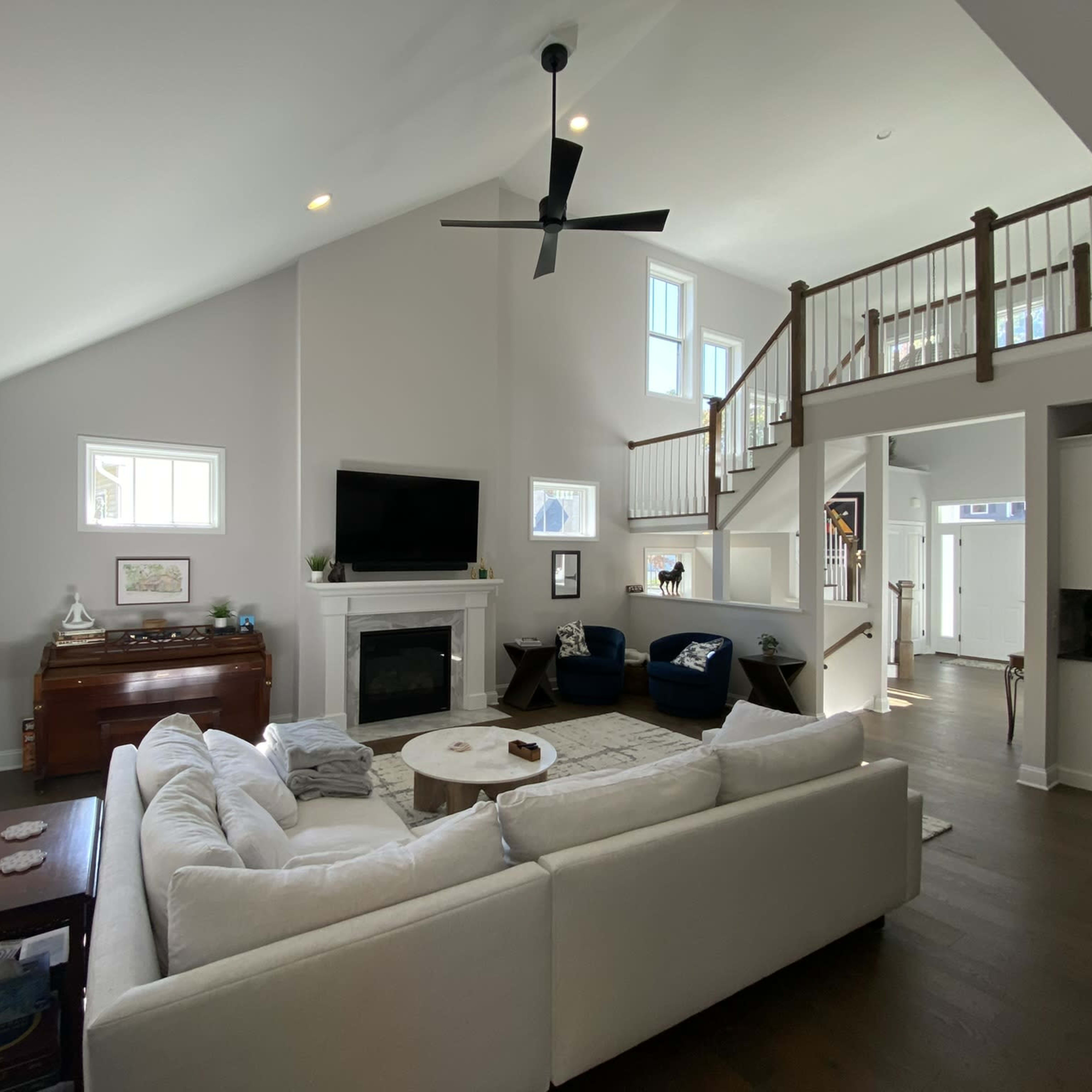 A spacious living room features a white sectional sofa, a large television above a fireplace, and wooden stairs leading to an upper level.