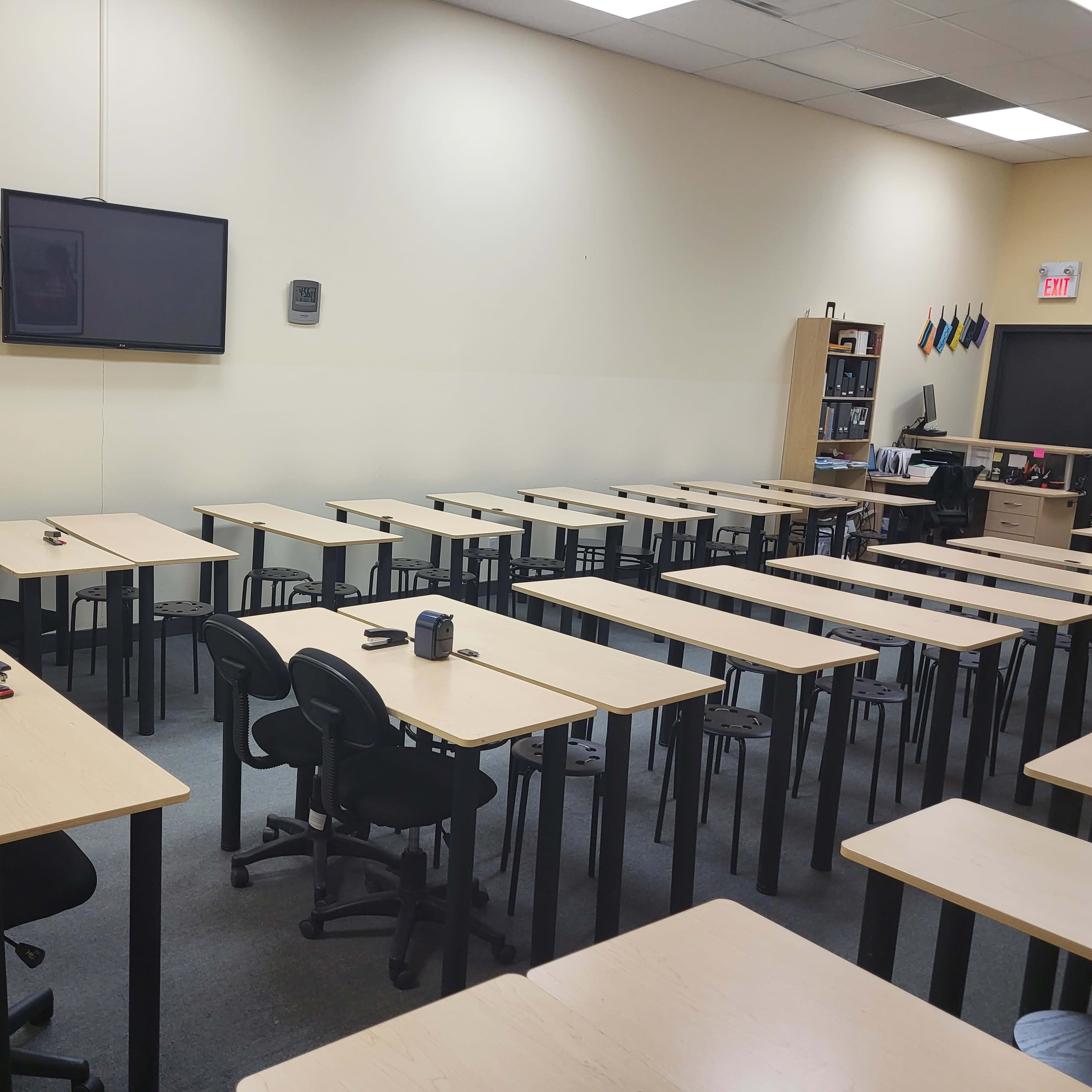 A classroom is set up with rows of tables and chairs, featuring a wall-mounted screen and a shelf with various items in the background.