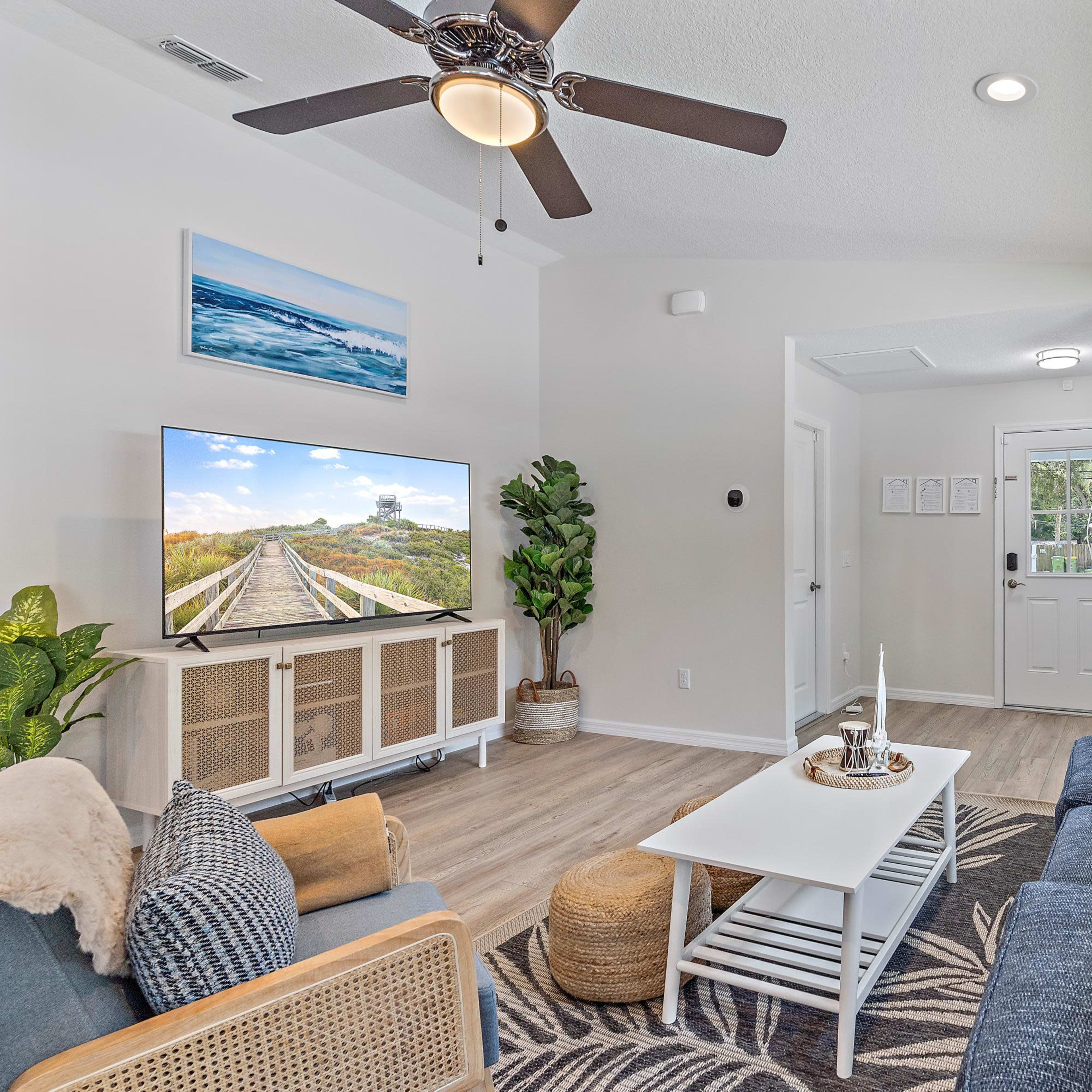 A bright living room with a ceiling fan, a large television, a coffee table, and comfortable seating, along with a doorway leading outside.
