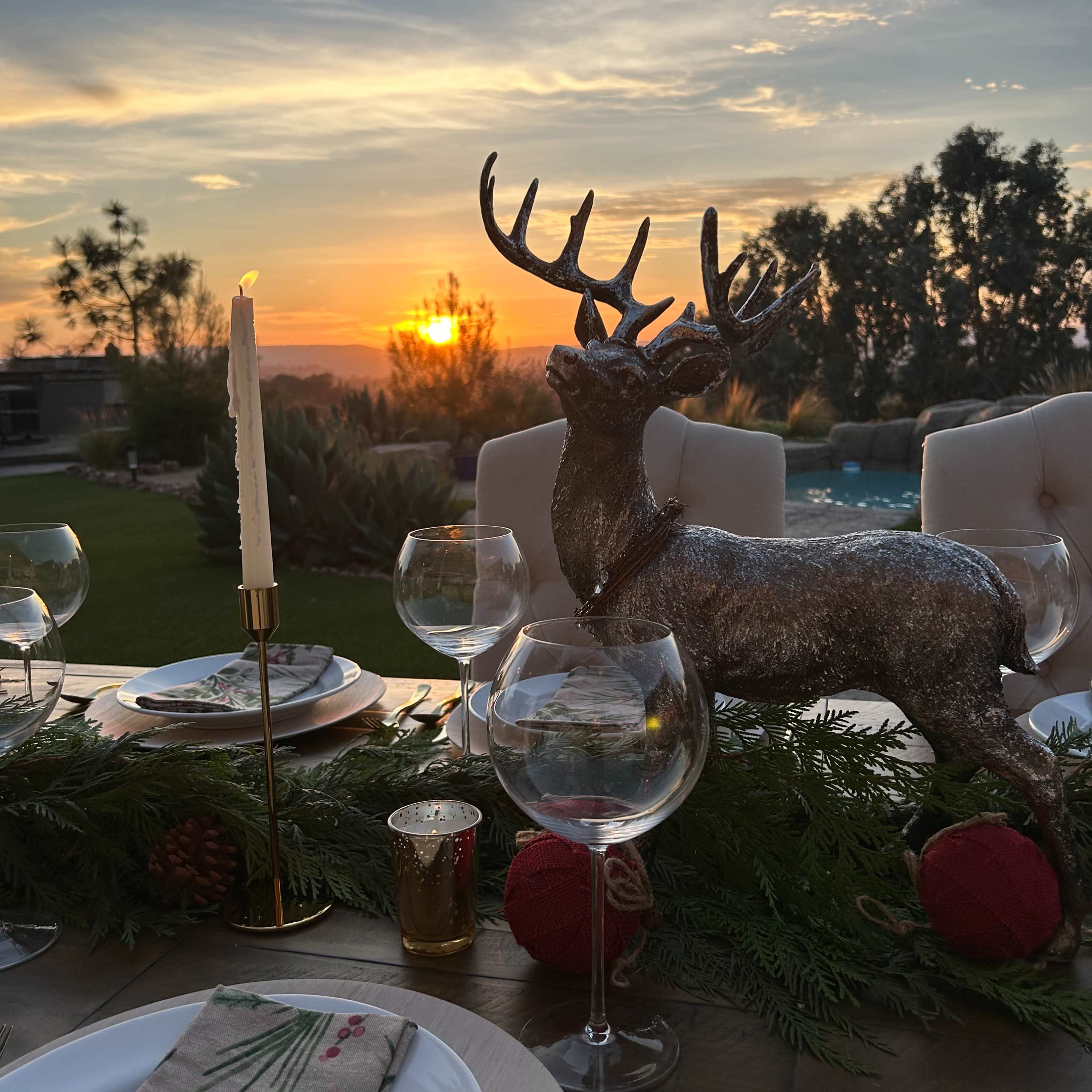 A festive table setting with a decorative reindeer centerpiece, surrounded by wine glasses and green garlands, overlooks a sunset by a pool.