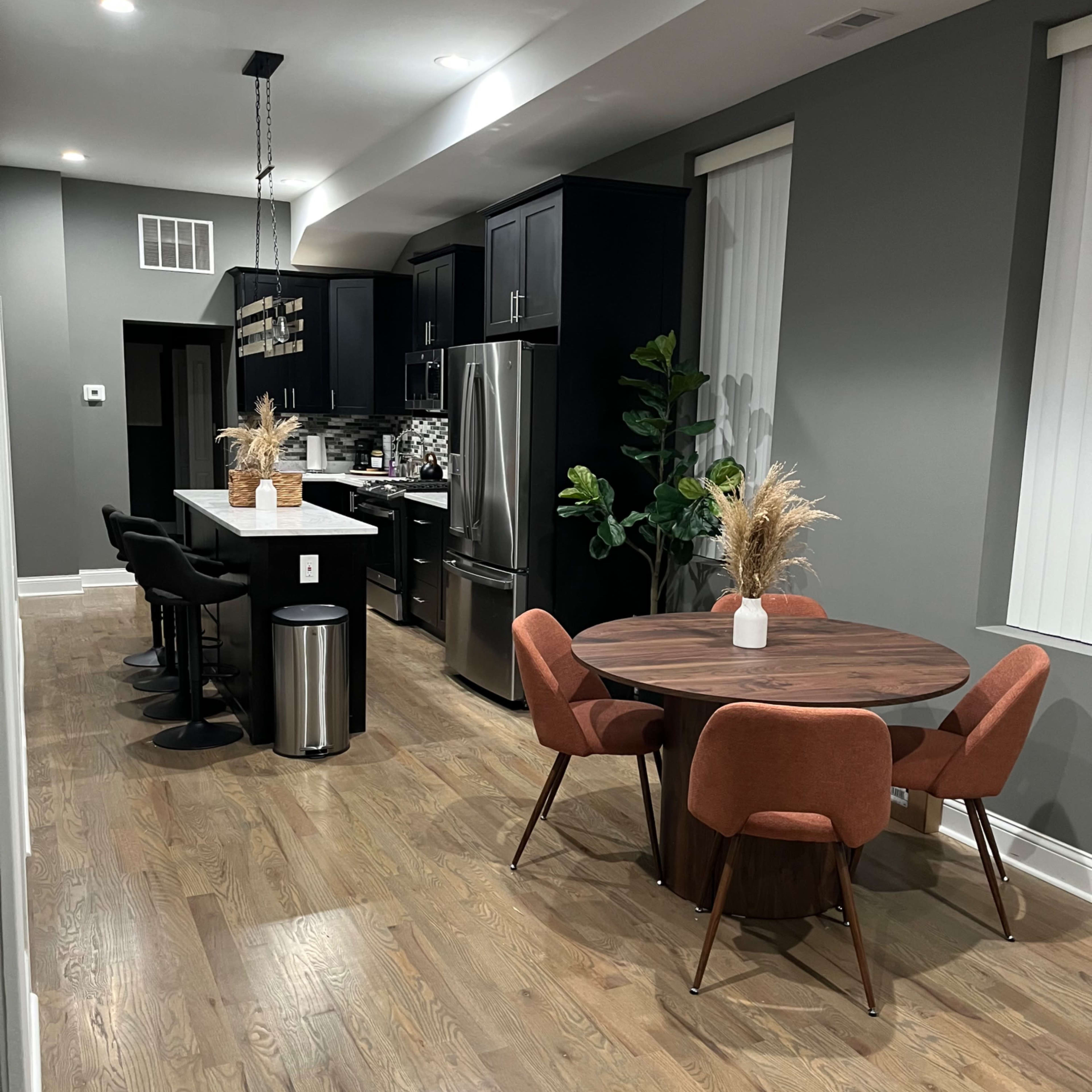 A modern kitchen and dining area featuring black cabinetry, stainless steel appliances, a circular wooden dining table with orange chairs, and a bar with black stools.