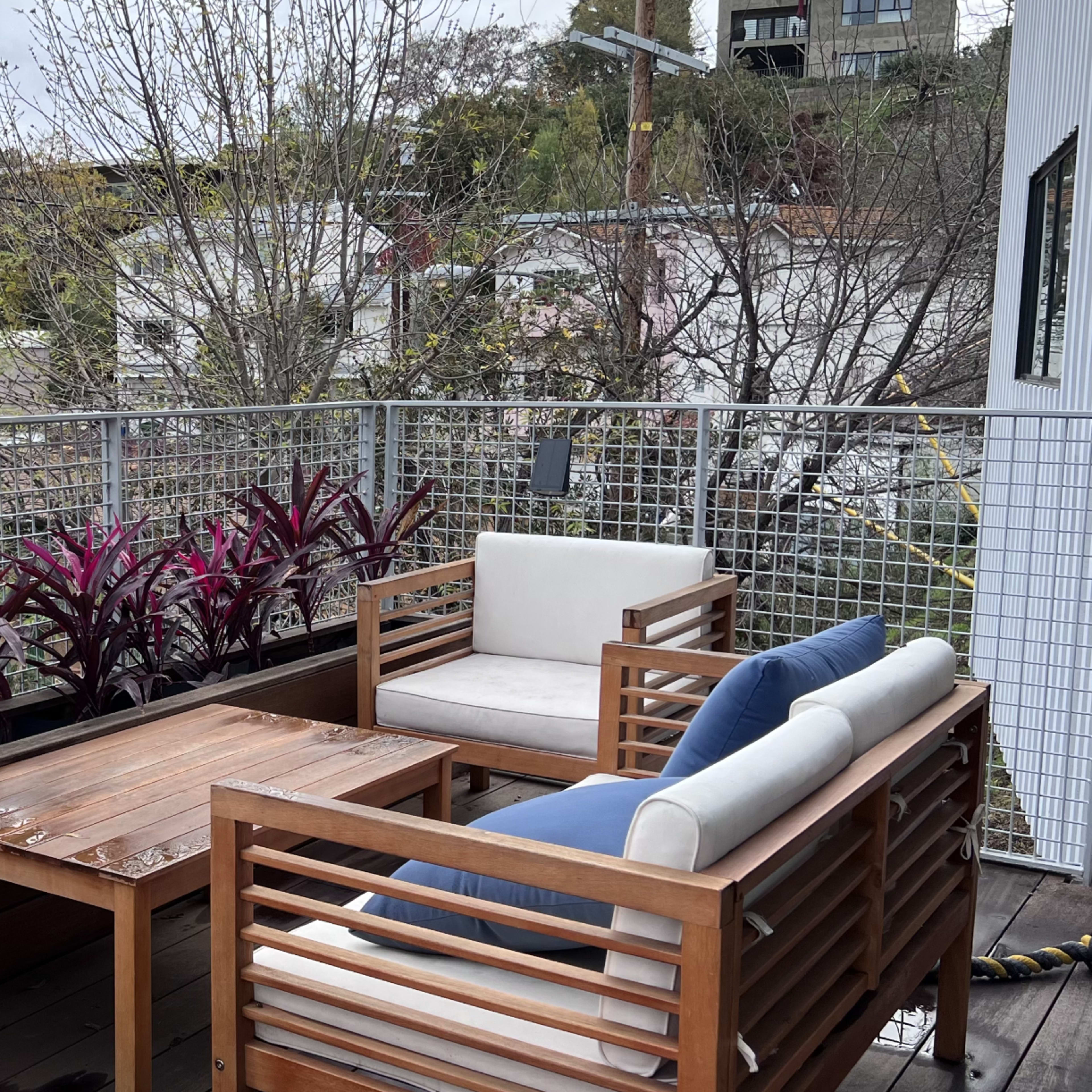 A wooden patio set with white cushions is arranged around a table, surrounded by greenery and purple plants on a balcony.