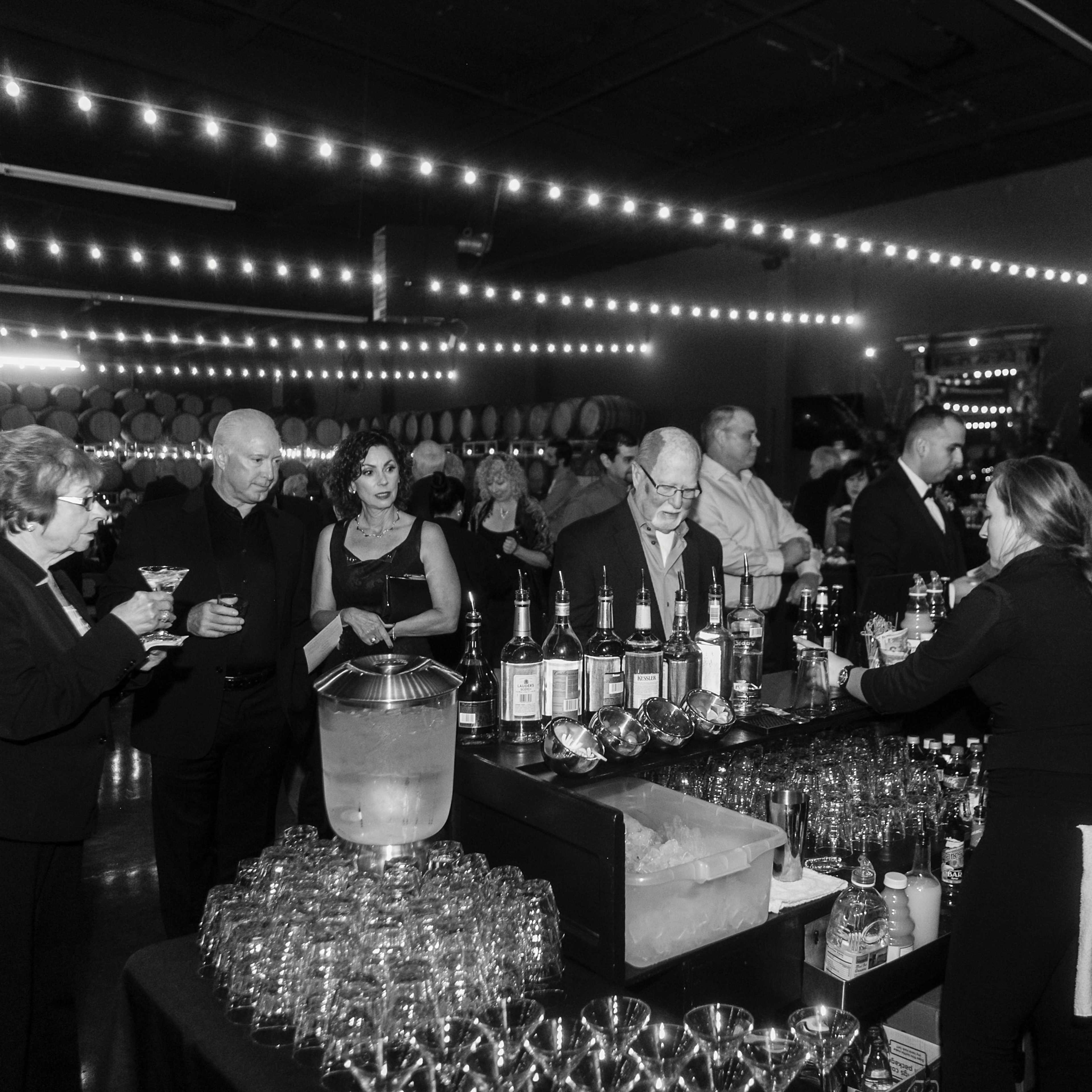 A group of people gathers at a bar with various drinks and an assortment of bottles in a dimly lit venue decorated with string lights.