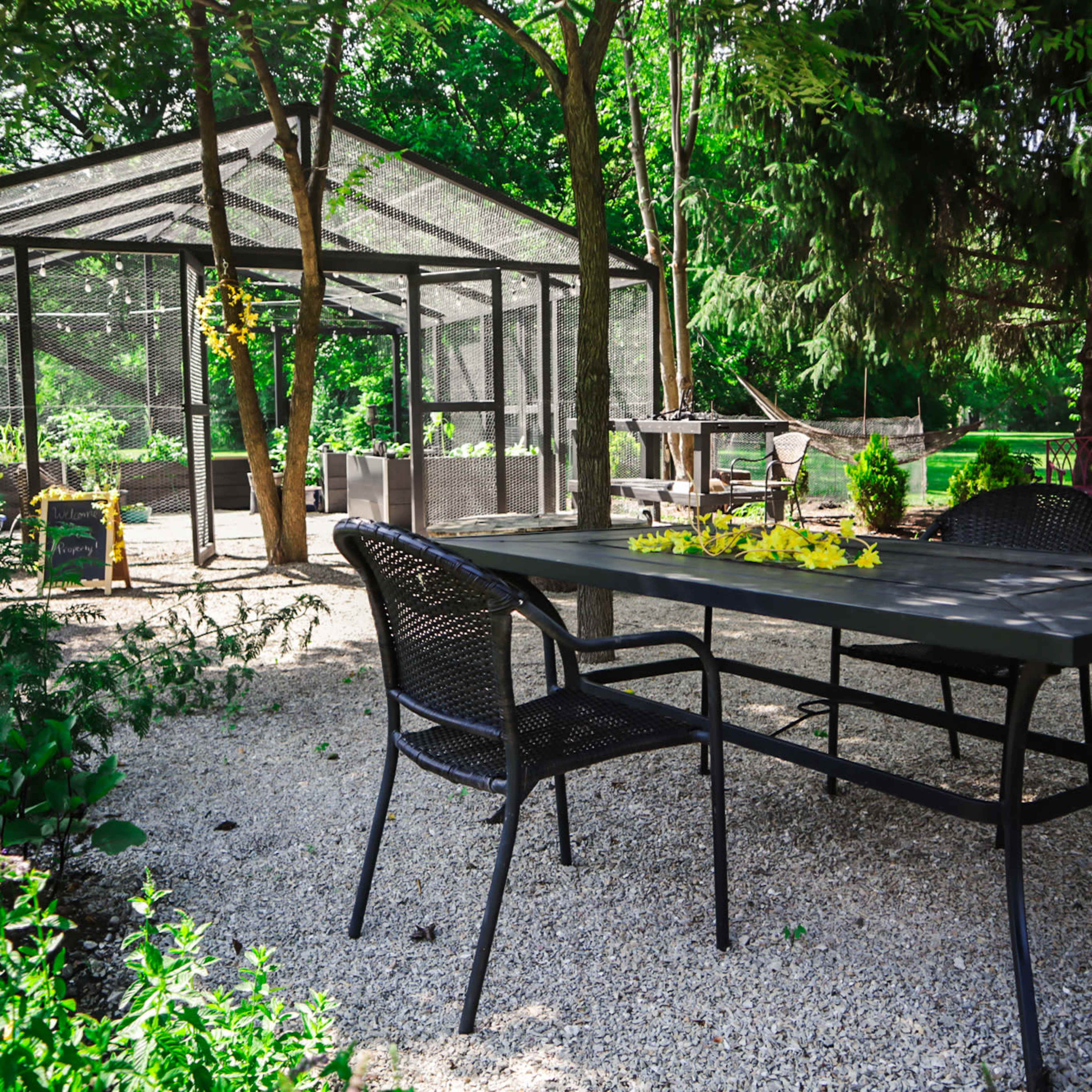 A metal table and chairs sit on gravel in a garden area near a glass greenhouse surrounded by greenery.