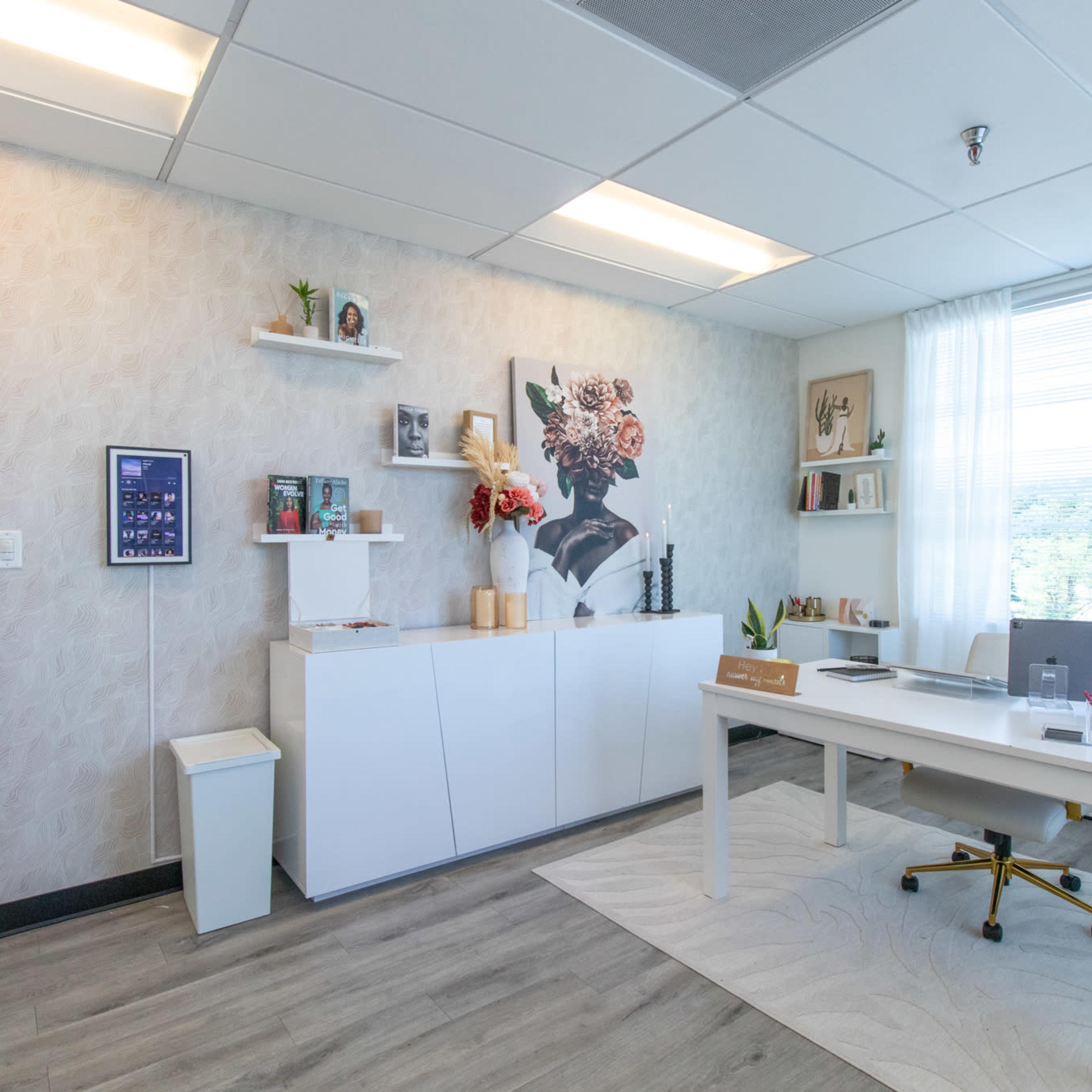 A well-organized office area features a white desk, a minimalistic shelf with decorative items, and a large window with sheer curtains letting in natural light.