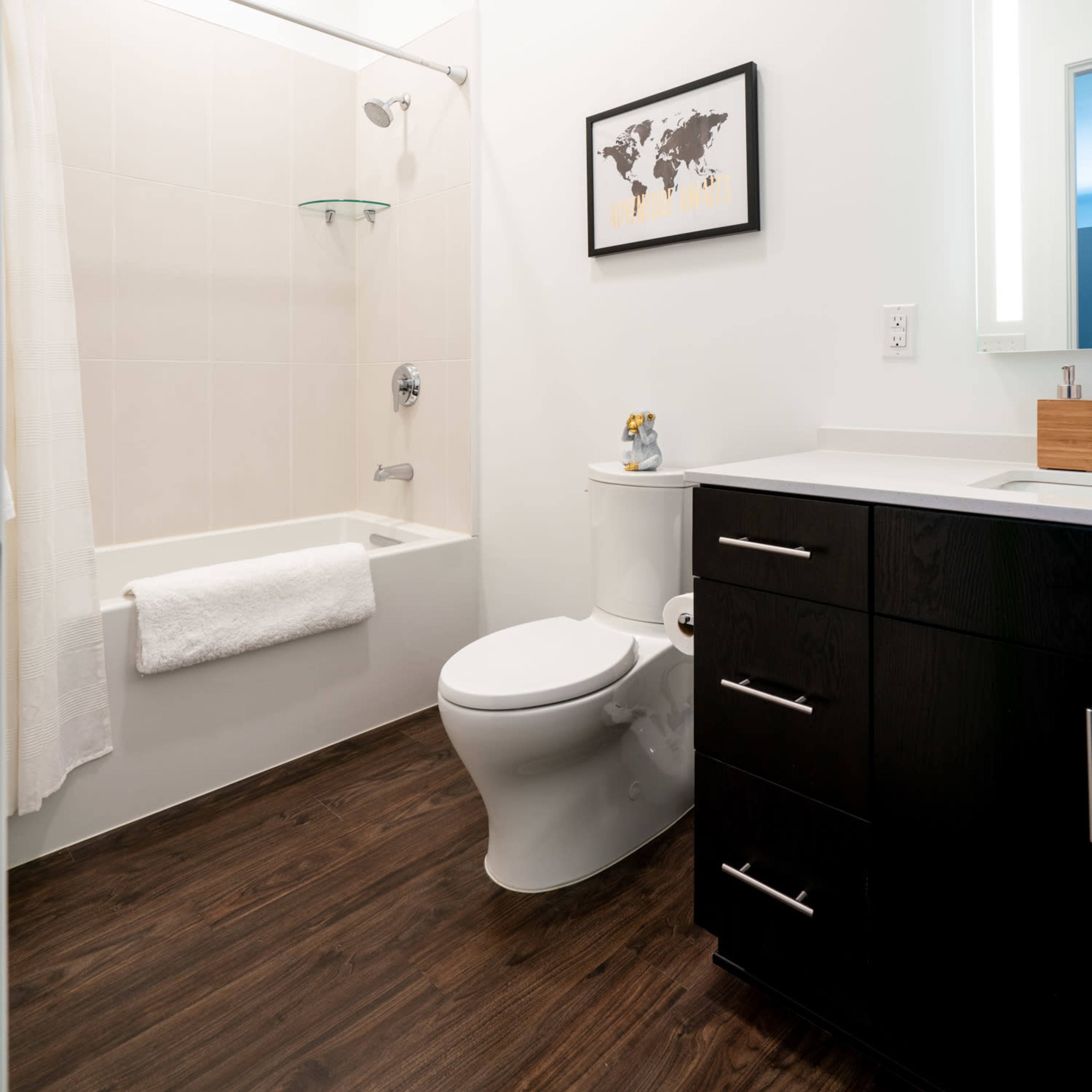 A modern bathroom with a bathtub, toilet, a dark cabinet with drawers, and a framed world map on the wall.