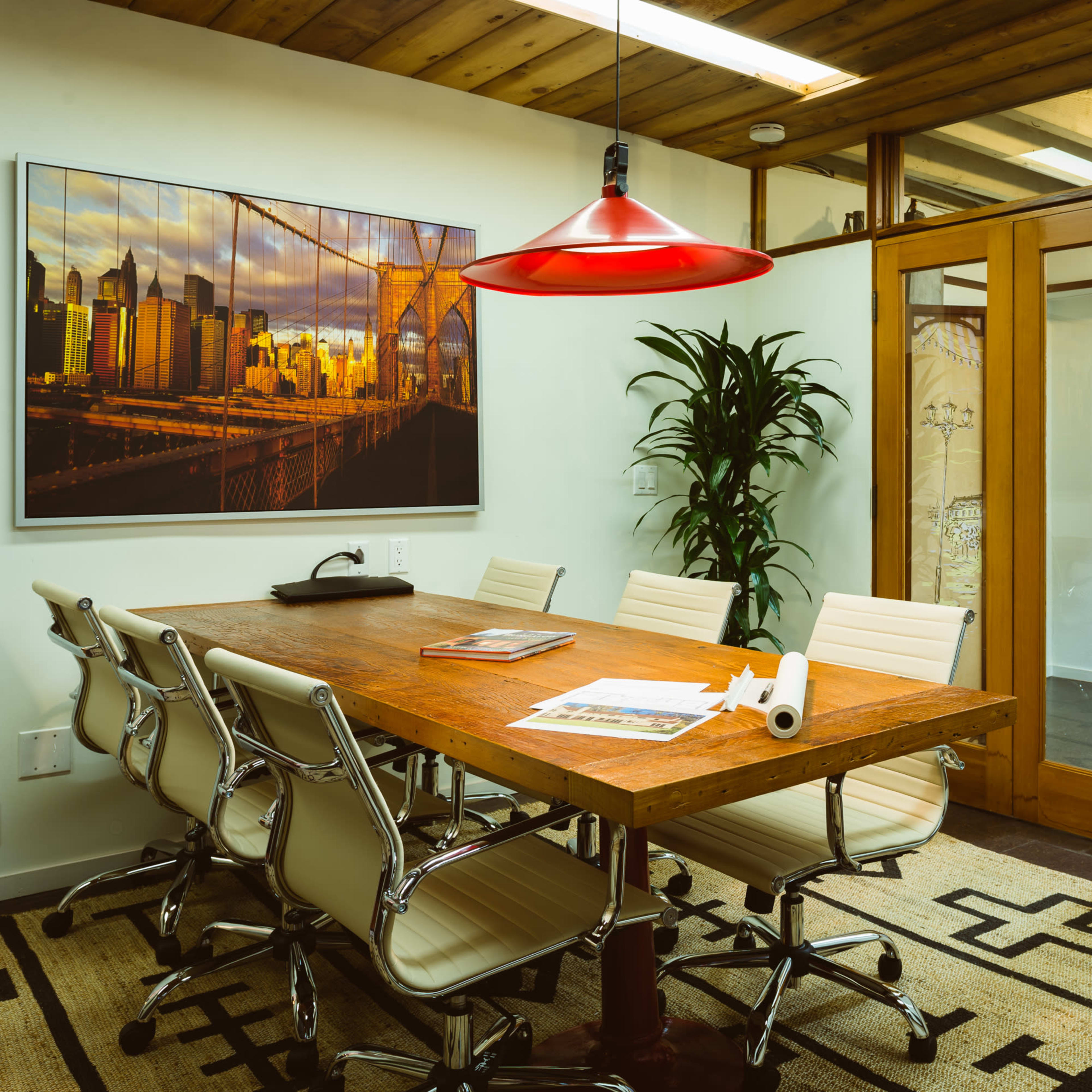 A modern conference room features a wooden table surrounded by white chairs, with a large artwork on the wall and a red pendant light overhead.