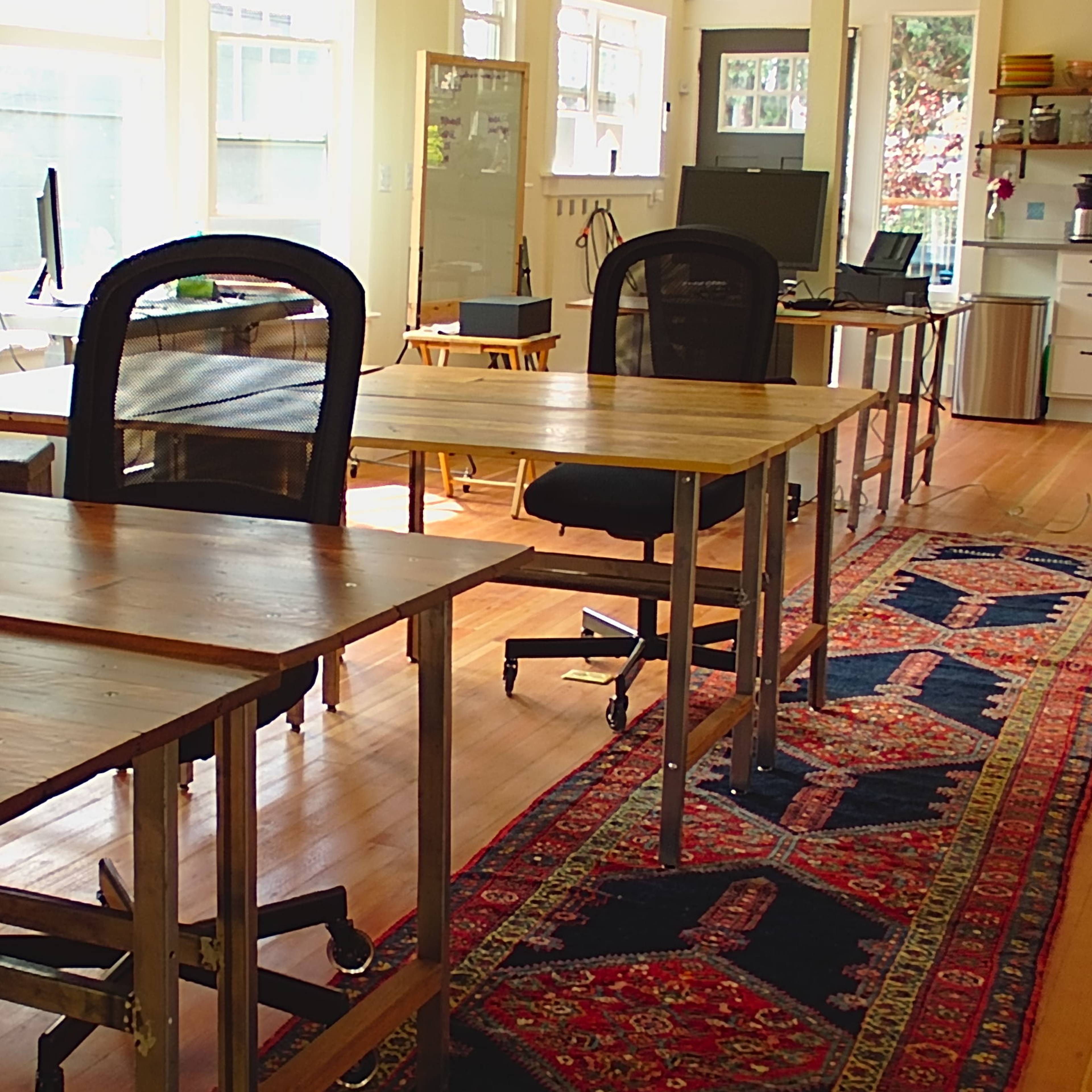 A bright room with several wooden tables and chairs arranged on a wooden floor, with a rug in the center and a kitchen area visible in the background.