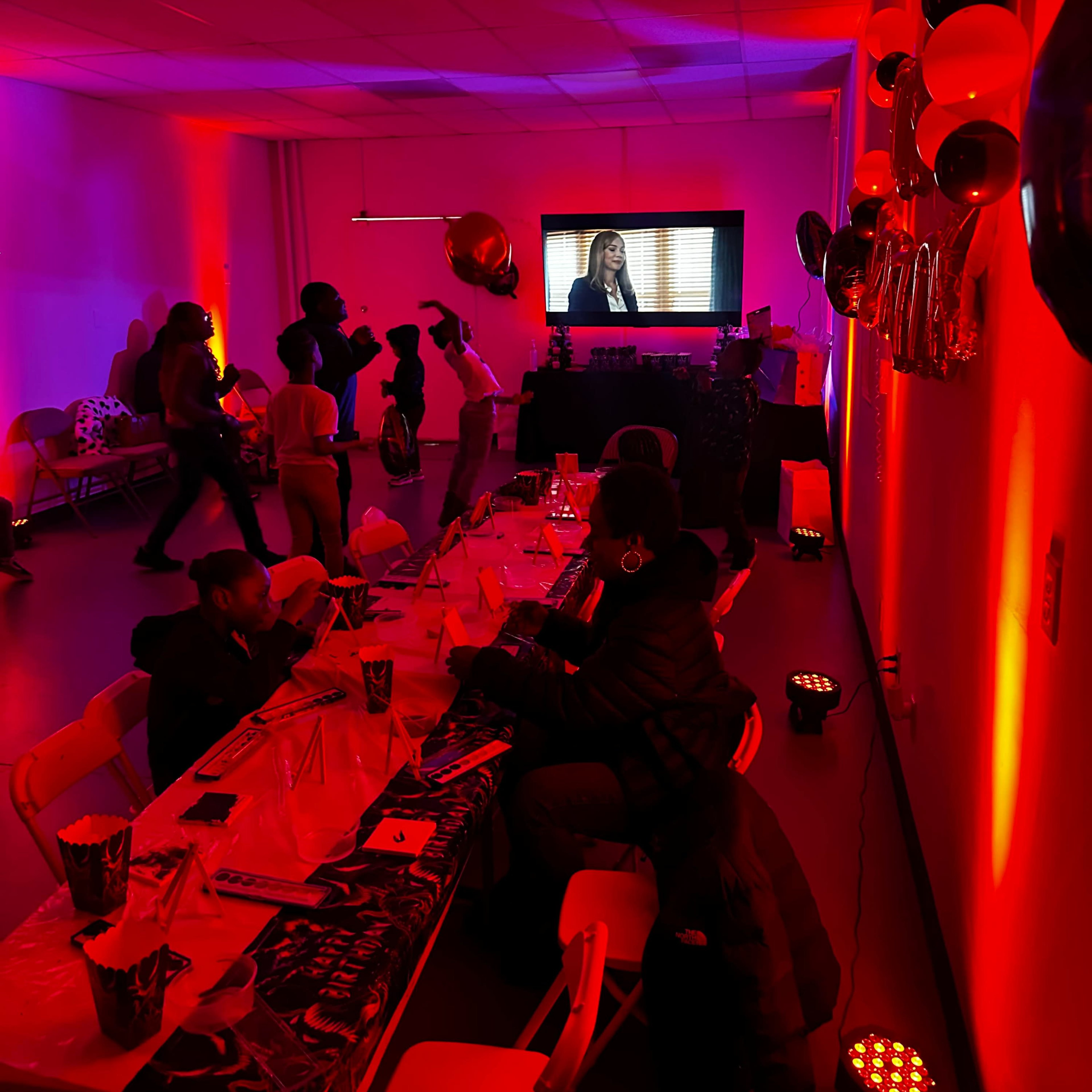 A gathering takes place in a dimly lit room with red lighting, featuring a table set for dining and a screen displaying a video.