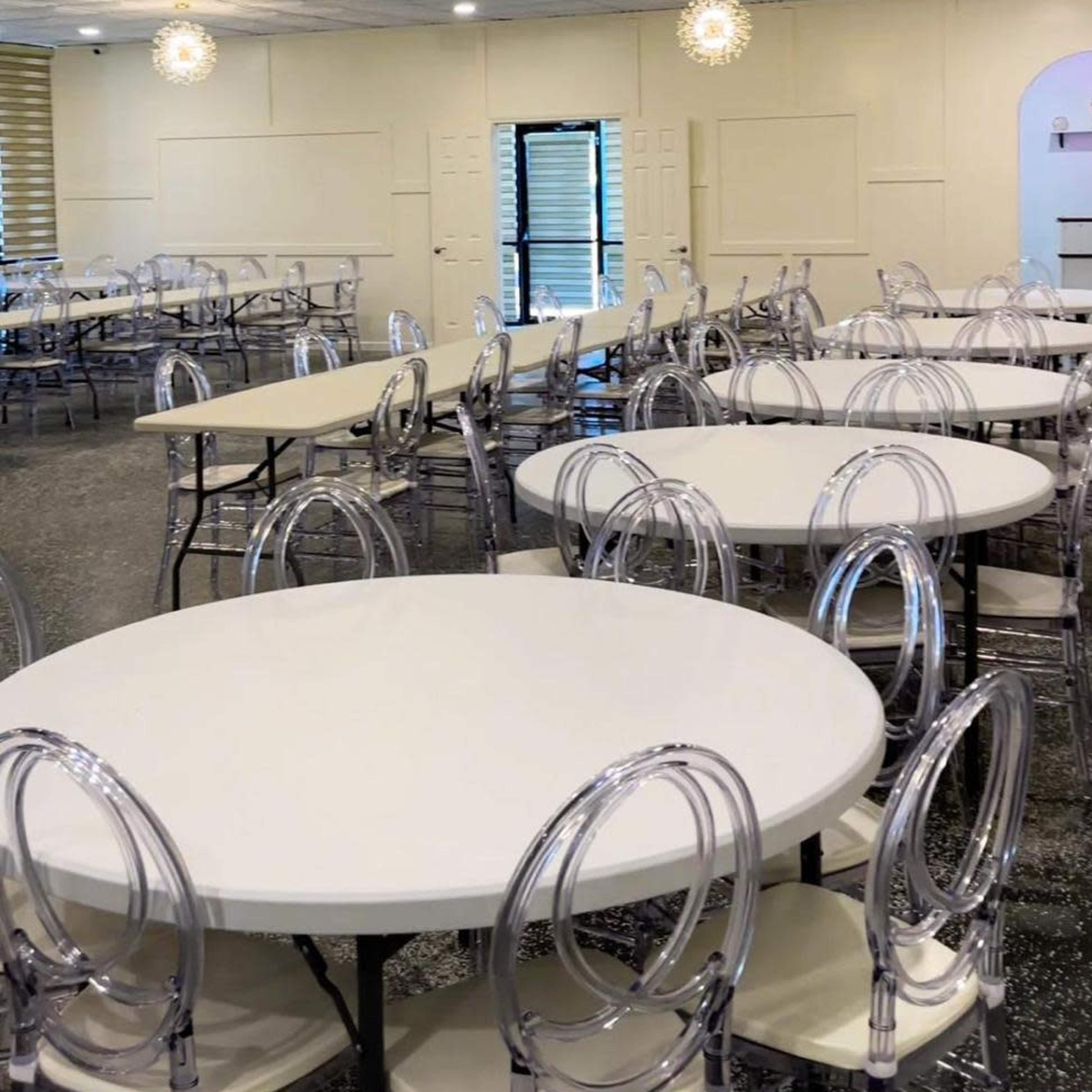 A spacious event room features round tables with clear chairs and pendant lighting on a textured ceiling.