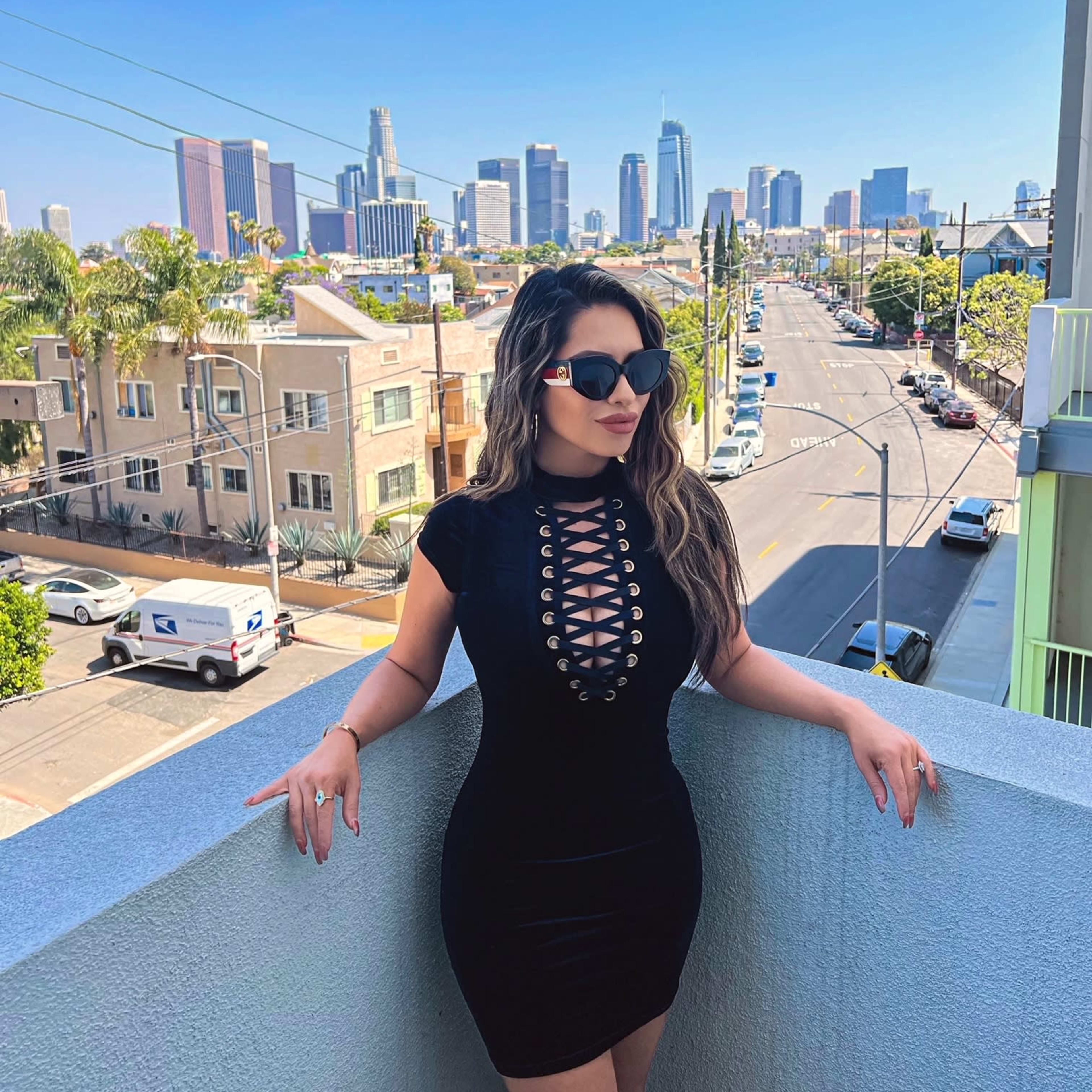 A woman in a form-fitting black dress stands on a balcony overlooking a city skyline with palm trees and streets visible below.