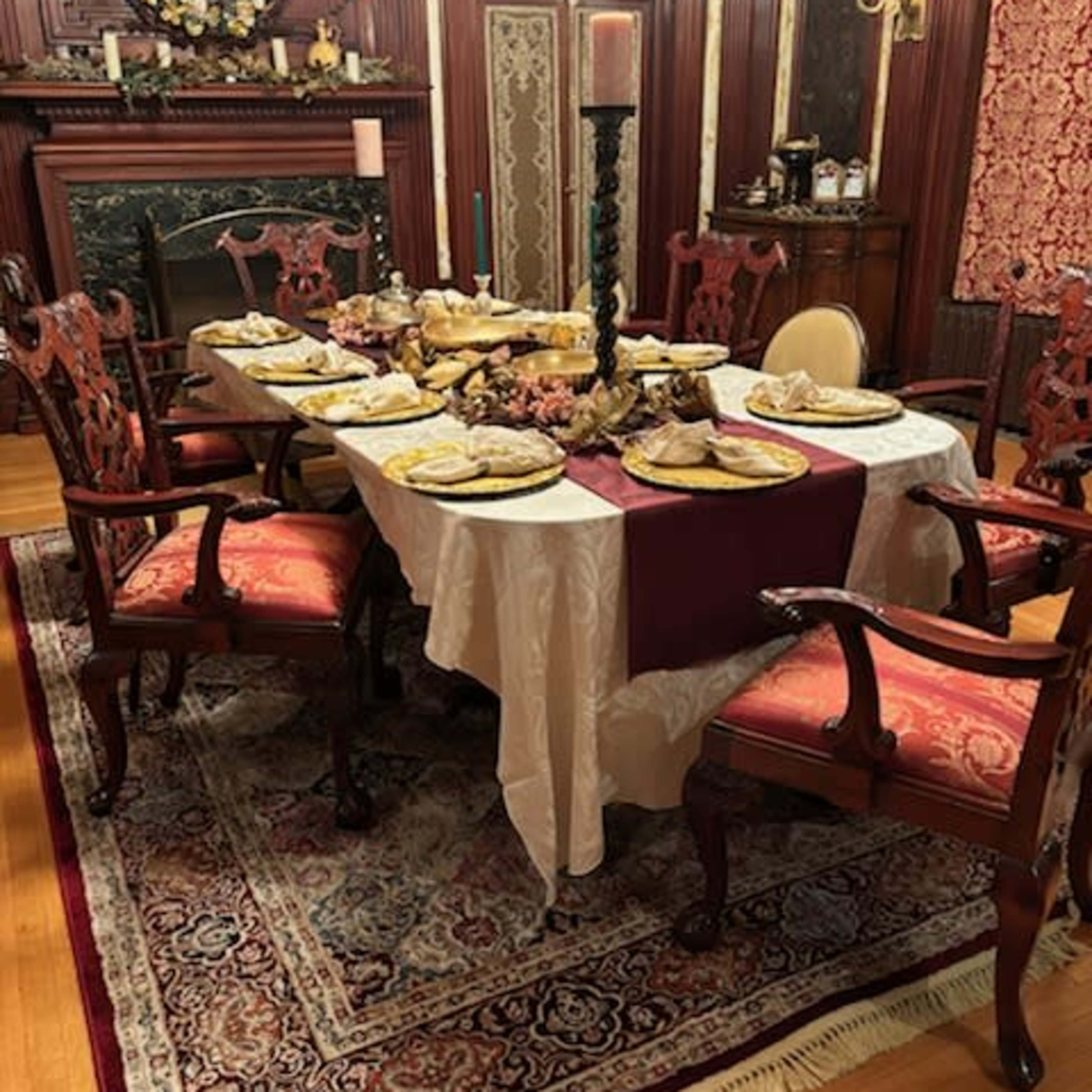 A dining table is set with multiple plates of food, surrounded by ornate chairs in a room with wooden paneling and a decorative fireplace.
