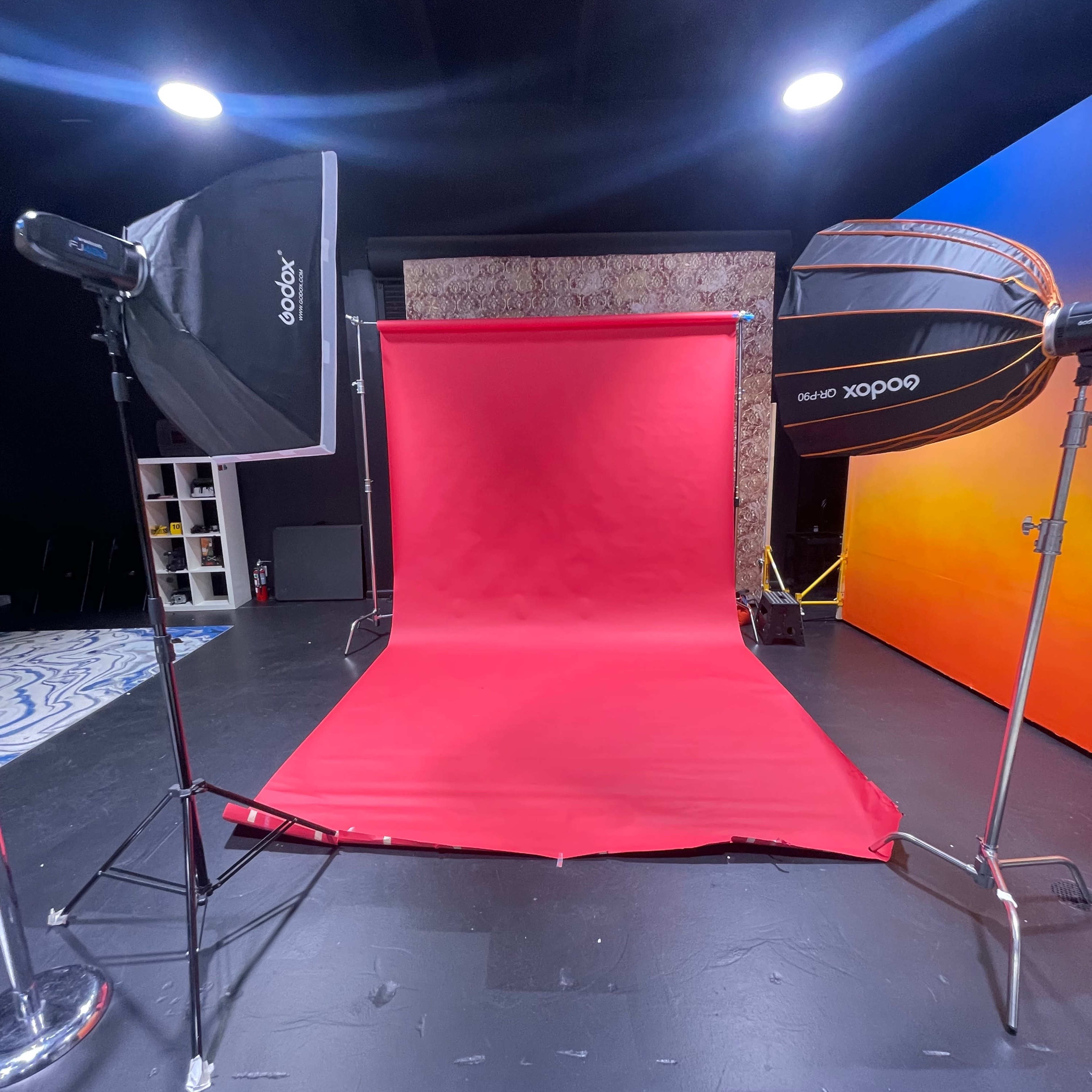 A photography studio setup featuring a red backdrop, two softbox lights, and a patterned wall in the background.