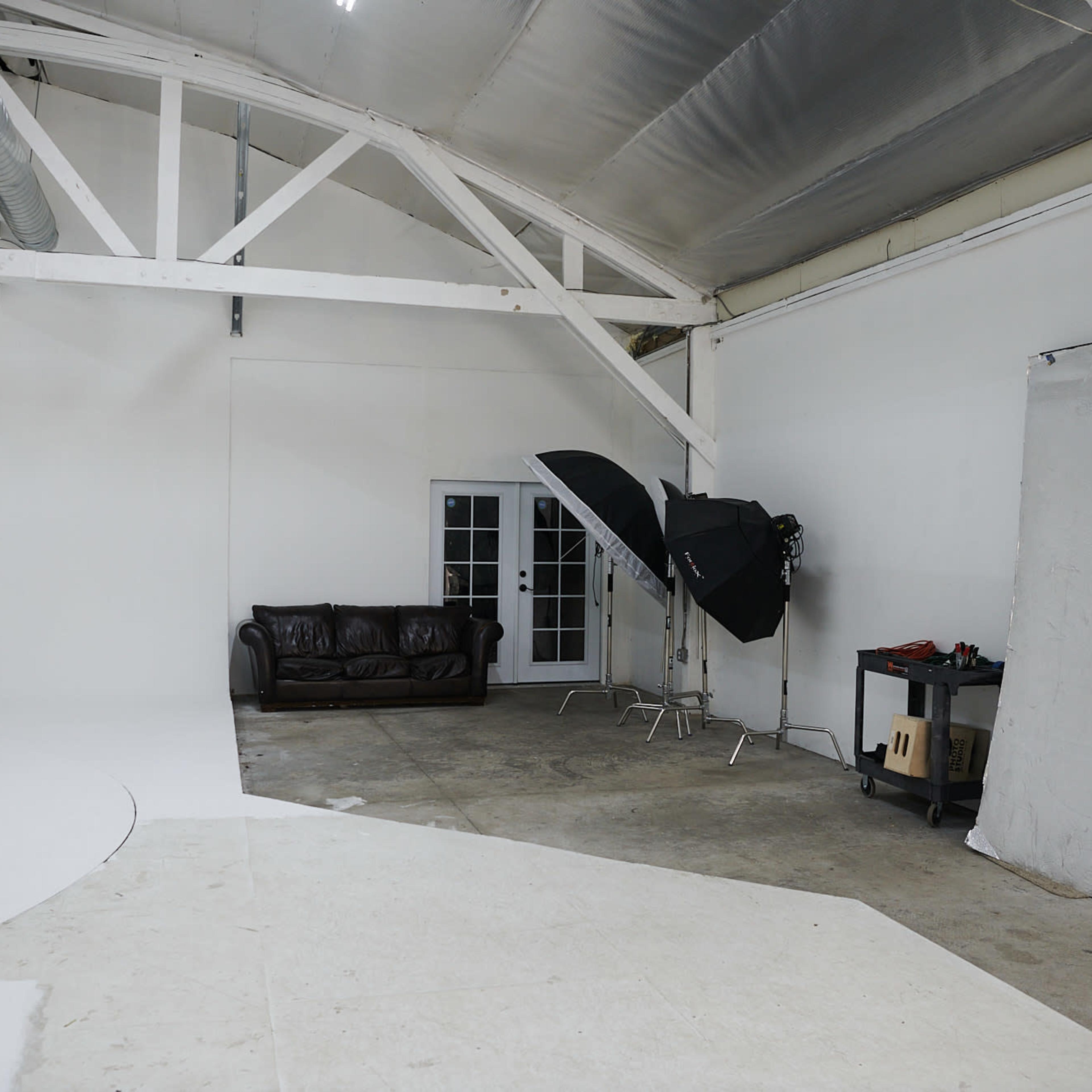 A spacious photography studio with a white backdrop, a leather couch, lighting equipment, and a small table against the wall.