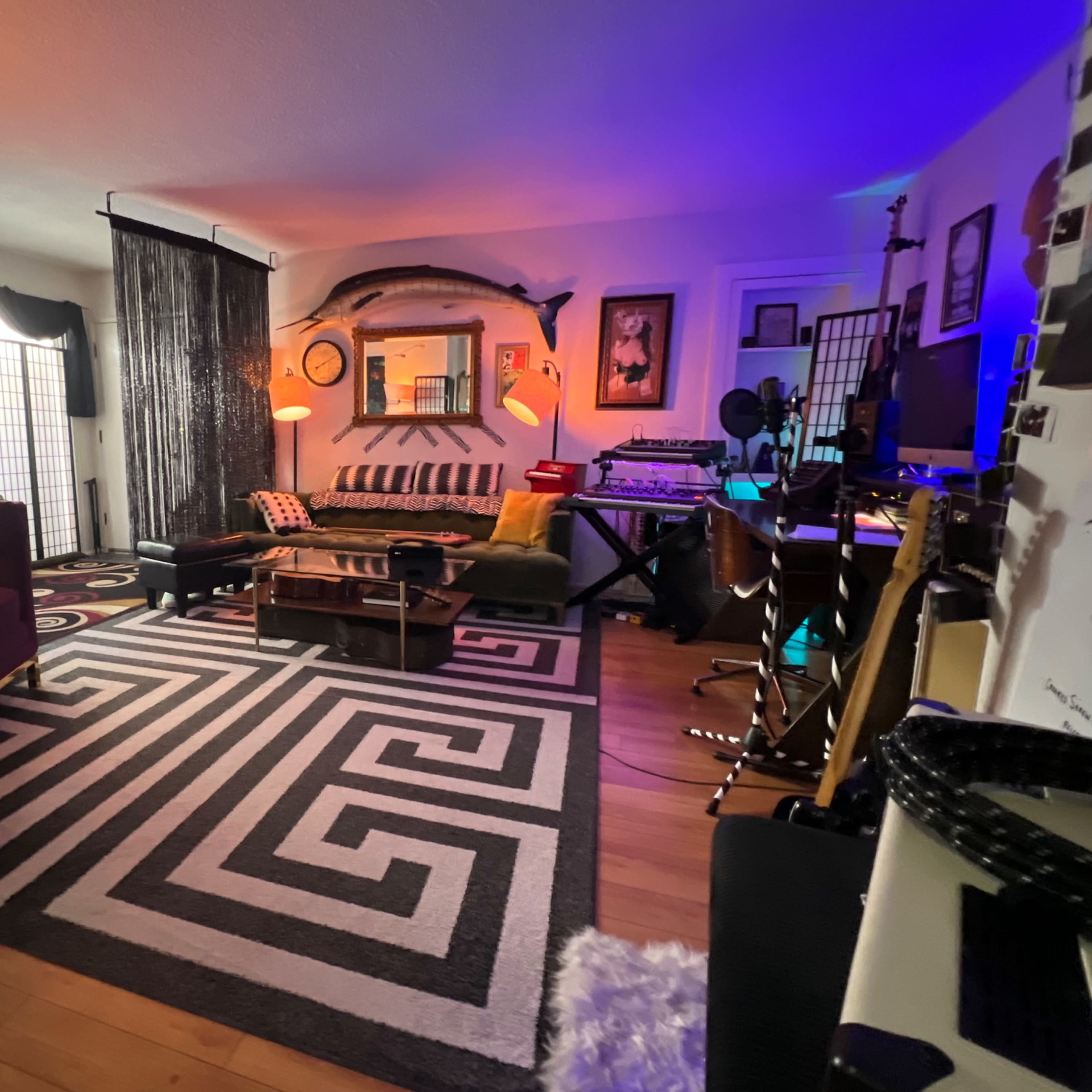The room features a patterned black and white rug, various seating areas, plants, and a mix of musical instruments and electronic equipment.