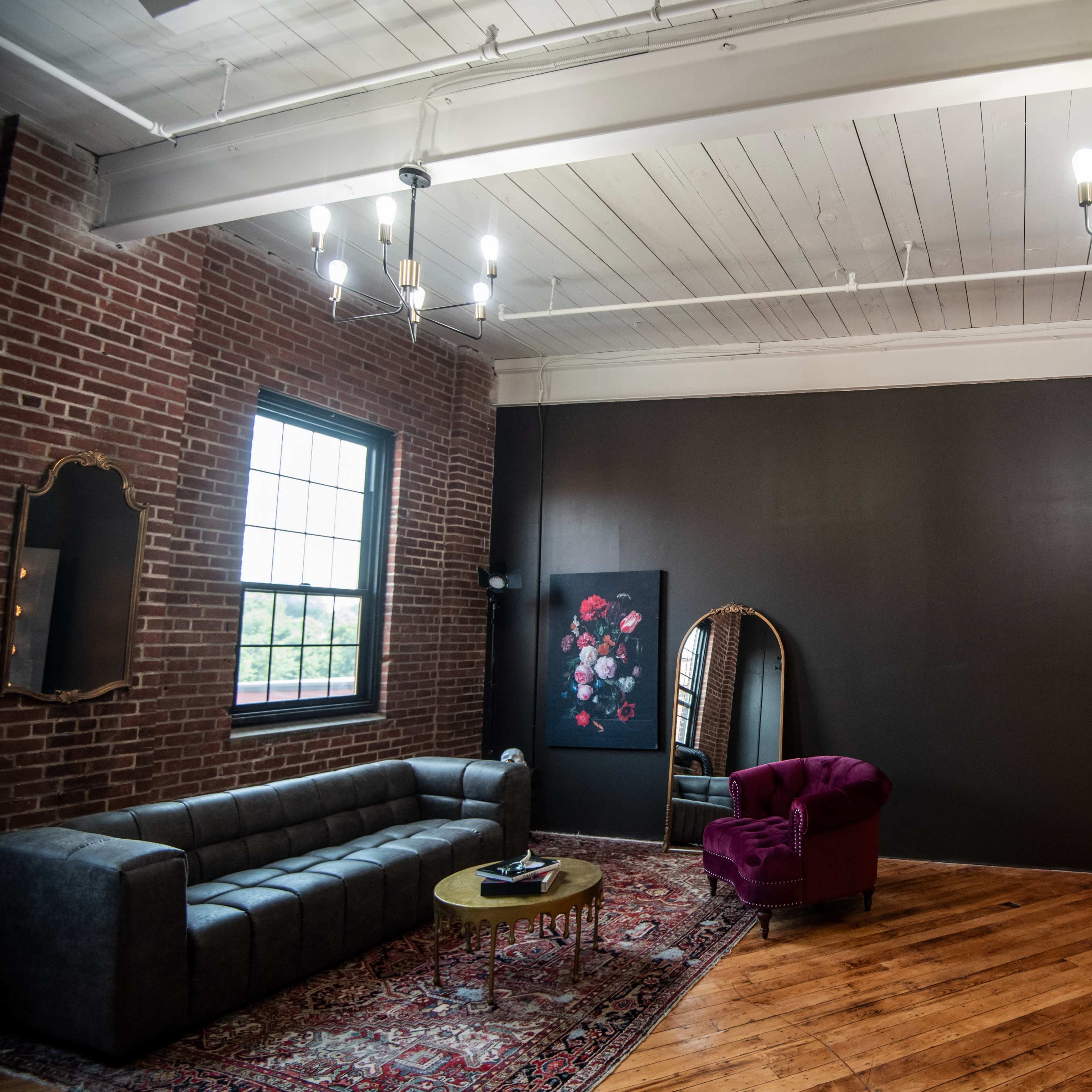 A spacious living area featuring a gray sofa, a round table, a purple chair, and a decorative wall with lights, against a backdrop of exposed brick and dark walls.