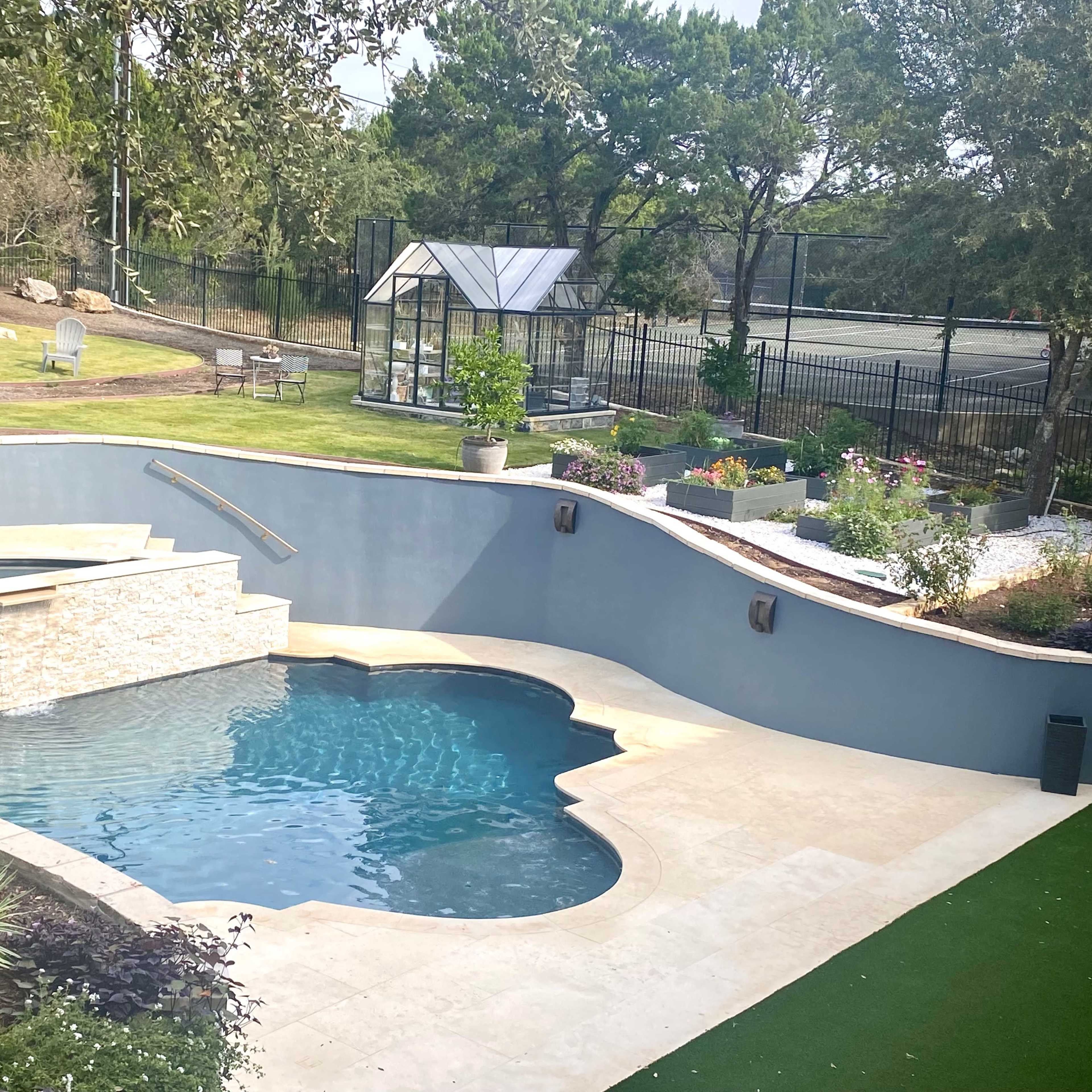 A backyard with a swimming pool featuring a stone wall, landscaped greenery, and a greenhouse in the background.