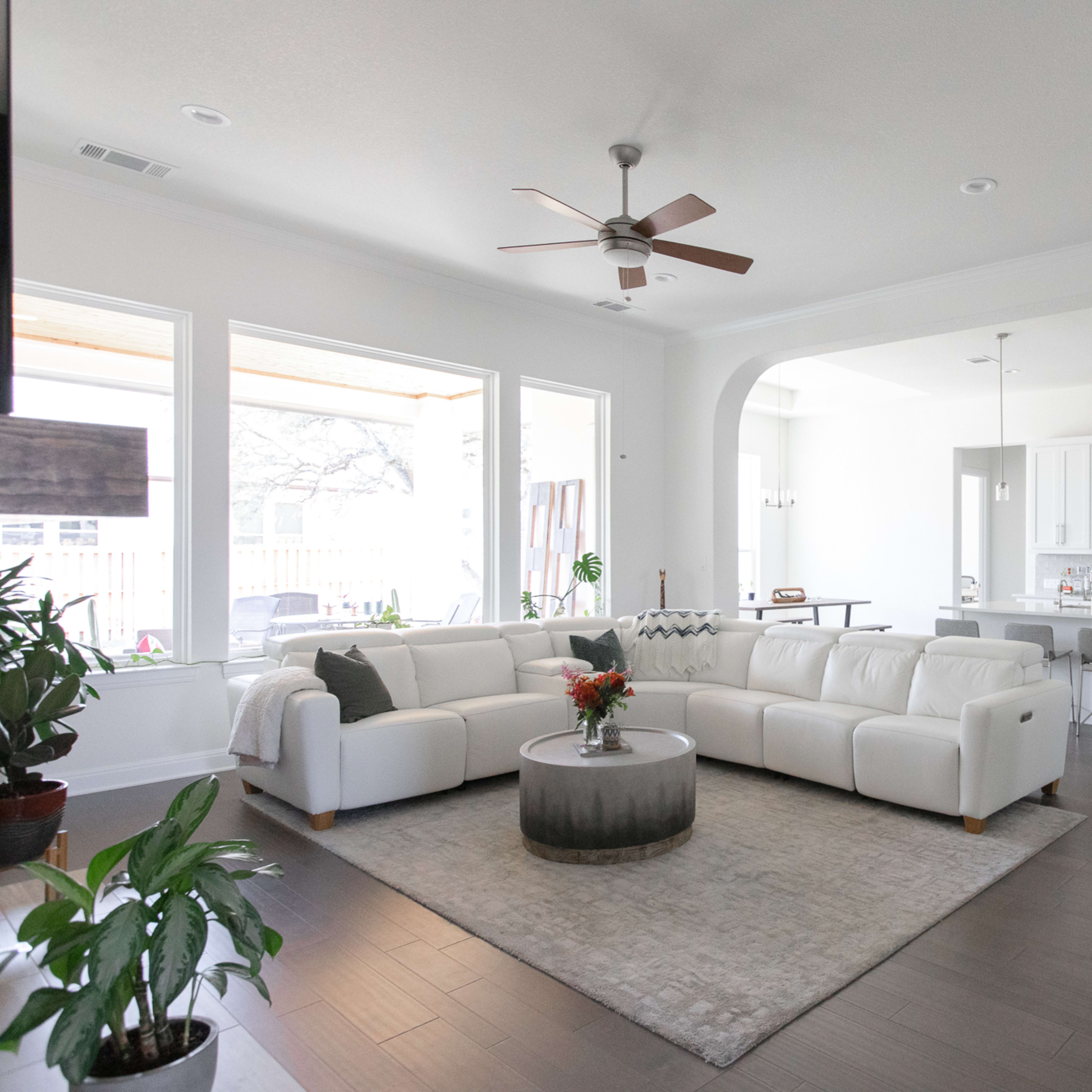 A spacious living room features a white sectional sofa, a round coffee table, and a view of an adjacent kitchen with modern appliances and fixtures.