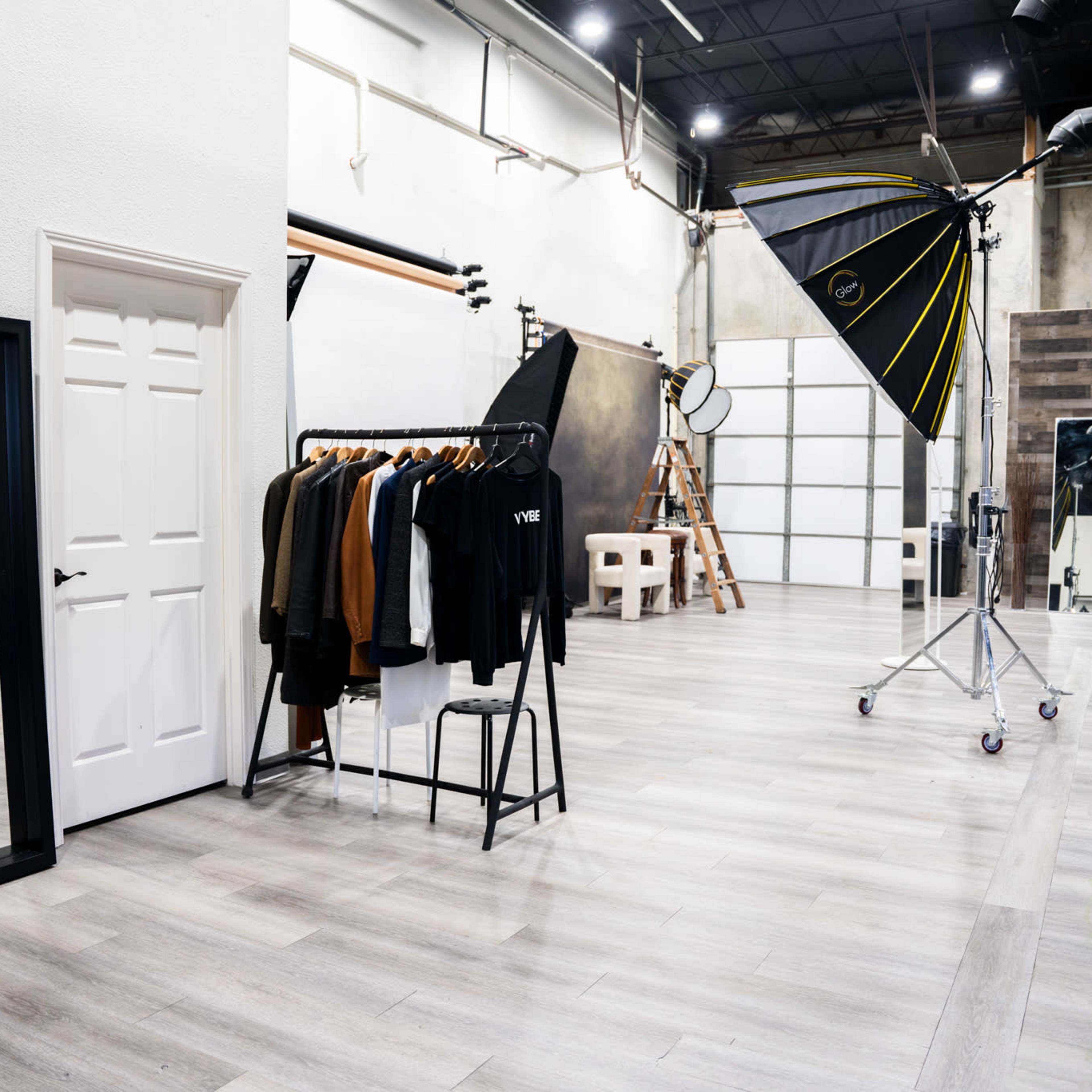 A spacious photography studio featuring a clothing rack, a large mirror, lighting equipment, and a backdrop area.