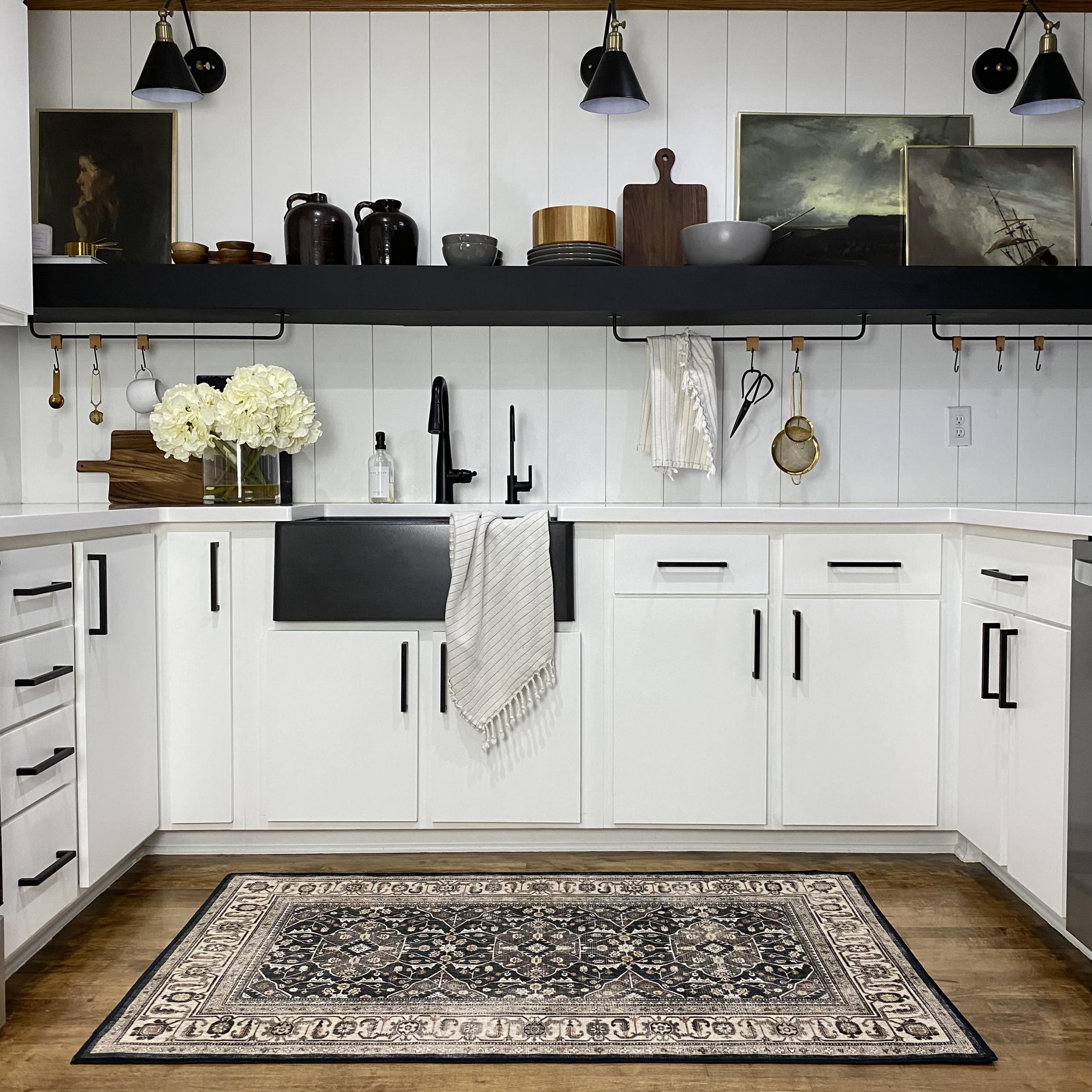 A modern kitchen features white cabinetry, a black farmhouse sink, and decorative items on a black shelf, with a vintage-style rug on the wooden floor.