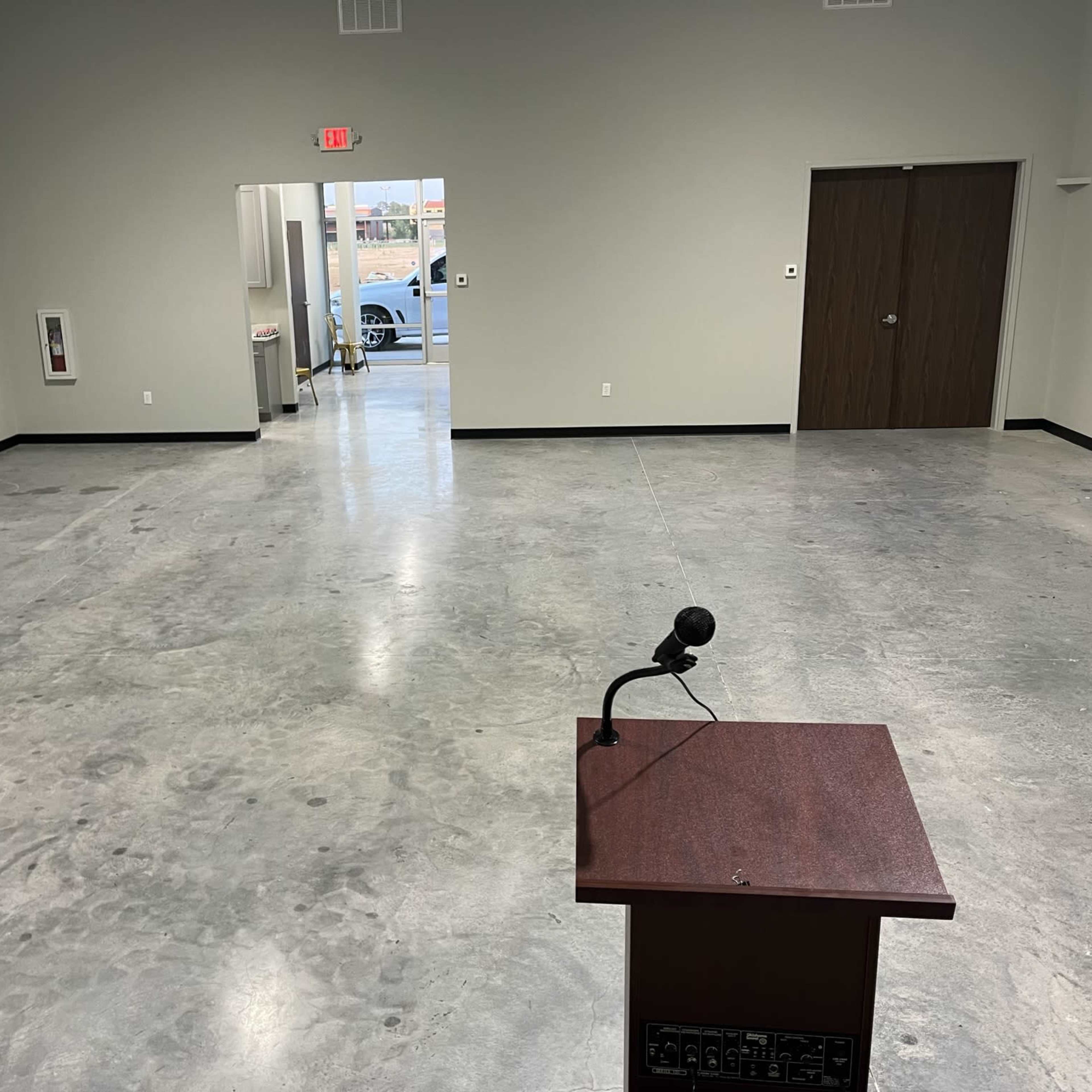 An empty room with a podium and microphone at the front, featuring a polished concrete floor and two doors leading to other areas.