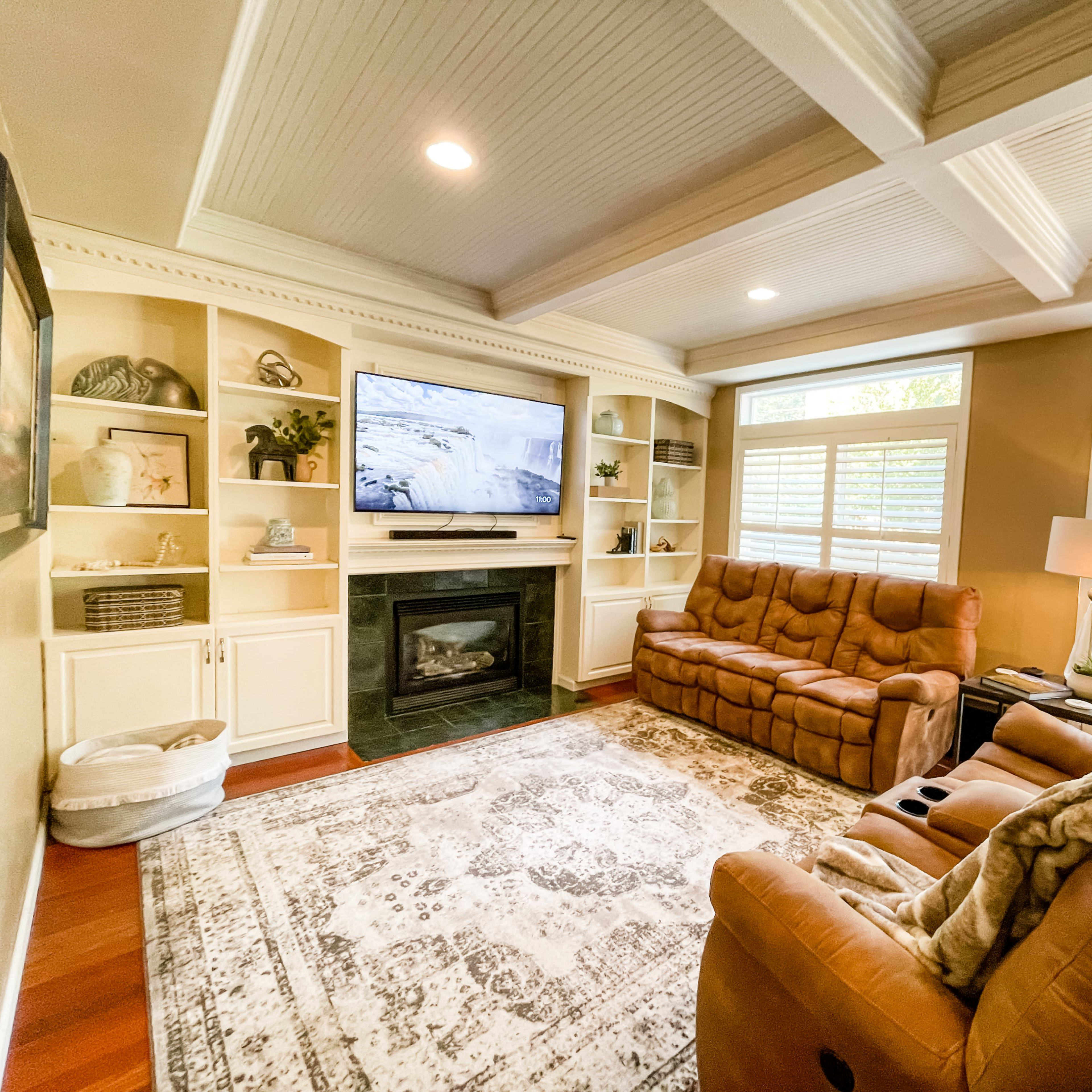 A cozy living room with a brown reclining sofa, a television mounted above a black fireplace, built-in shelves, and large windows allowing natural light.